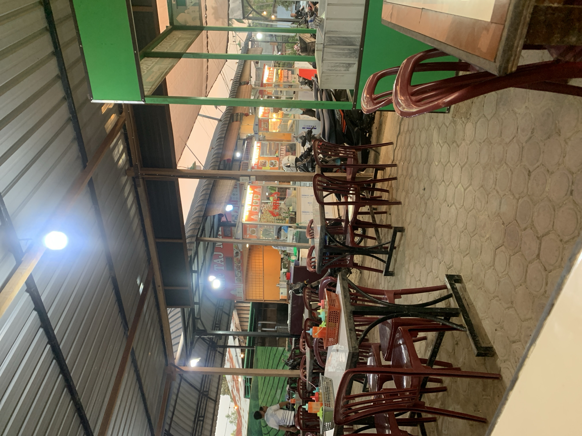An outdoor dining area with a metal roof supported by wooden beams is shown. The floor is paved with hexagonal interlocking concrete tiles. Red plastic chairs surround square and rectangular tables, some of which are set with condiment bottles and baskets. In the background, food stalls with illuminated signs display menus and food items. Several motorcycles are parked near the stalls. Two individuals are visible in the middle ground, one walking away from the camera and the other standing near a stall. The lighting suggests it is late afternoon or early evening.