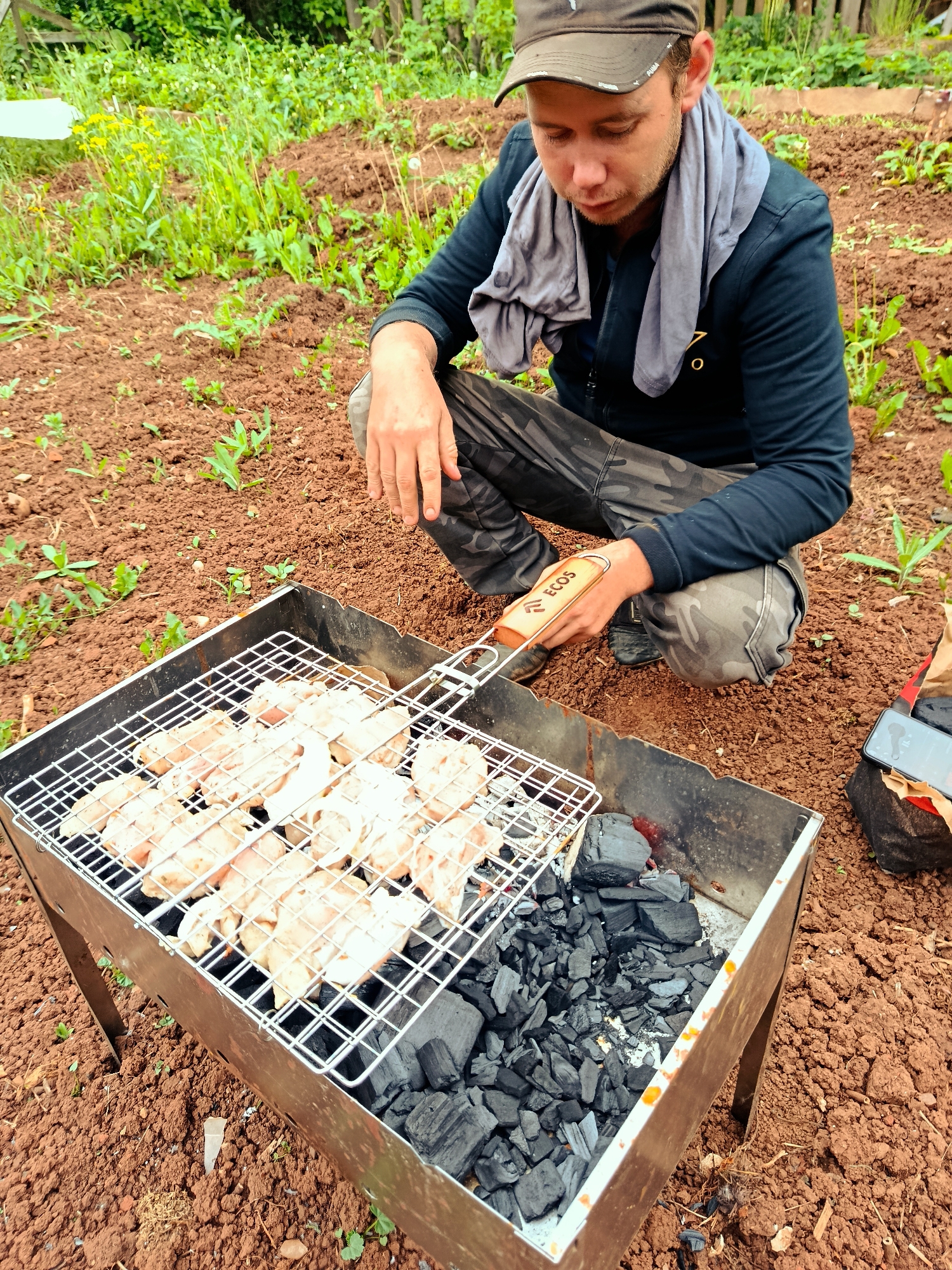 The image depicts an outdoor grilling scene in Фоки, Russia. A male individual, wearing a dark baseball cap, a grey t-shirt draped around his neck, a dark blue jacket, and camouflage trousers, is shown in a squatting position beside a rectangular metal charcoal grill. He holds a wooden-handled BBQ brush, branded "ECOS," and is actively applying a sauce or marinade to multiple pieces of raw or lightly seasoned chicken placed on a metal grate above burning charcoal briquettes. The ground consists of reddish-brown tilled earth with scattered young green plants and weeds. In the background, denser green foliage with some yellow flowers and a partial wooden fence are visible. A dark-colored smartphone with white earbuds rests on a piece of red fabric next to the grill on the right side of the frame.