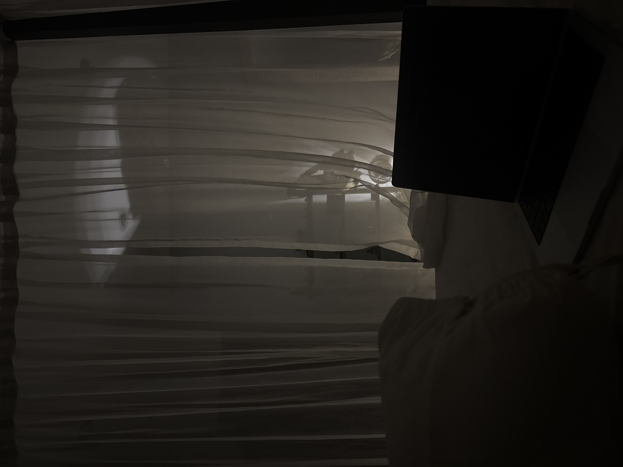 The image depicts a dimly lit bedroom scene, with sheer white curtains covering a window. Through the translucent fabric, the silhouette of a person can be vaguely seen, suggesting a presence behind the curtains. The overall ambiance is subdued and quiet, possibly indicating nighttime or early morning. A laptop, closed and dark, rests on a surface to the right, hinting at a workspace or personal use within the room. The focus is on the interplay of light and shadow, with the curtains creating a soft, diffused effect. The setting appears to be a bedroom in Indonesia, given the location context of Kuta Selatan. No specific activity is clearly visible beyond the implied presence of someone behind the curtains and the stationary laptop. The mood is serene and introspective, with the limited visibility adding an element of mystery.