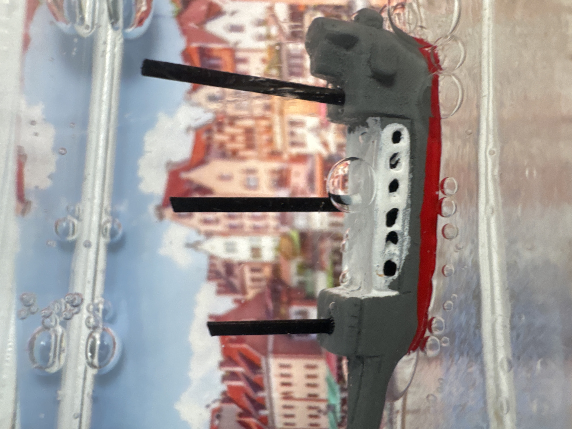 The image shows a model of a ship inside a clear, possibly liquid-filled container. The ship is gray with a red stripe along the side and features what appear to be gun barrels extending from its side. There are air bubbles surrounding the model and clinging to the container. The background is a blurred cityscape, depicting buildings with red tile roofs and light-colored walls. The scene looks like something in the city of Burgas, Bulgaria.
