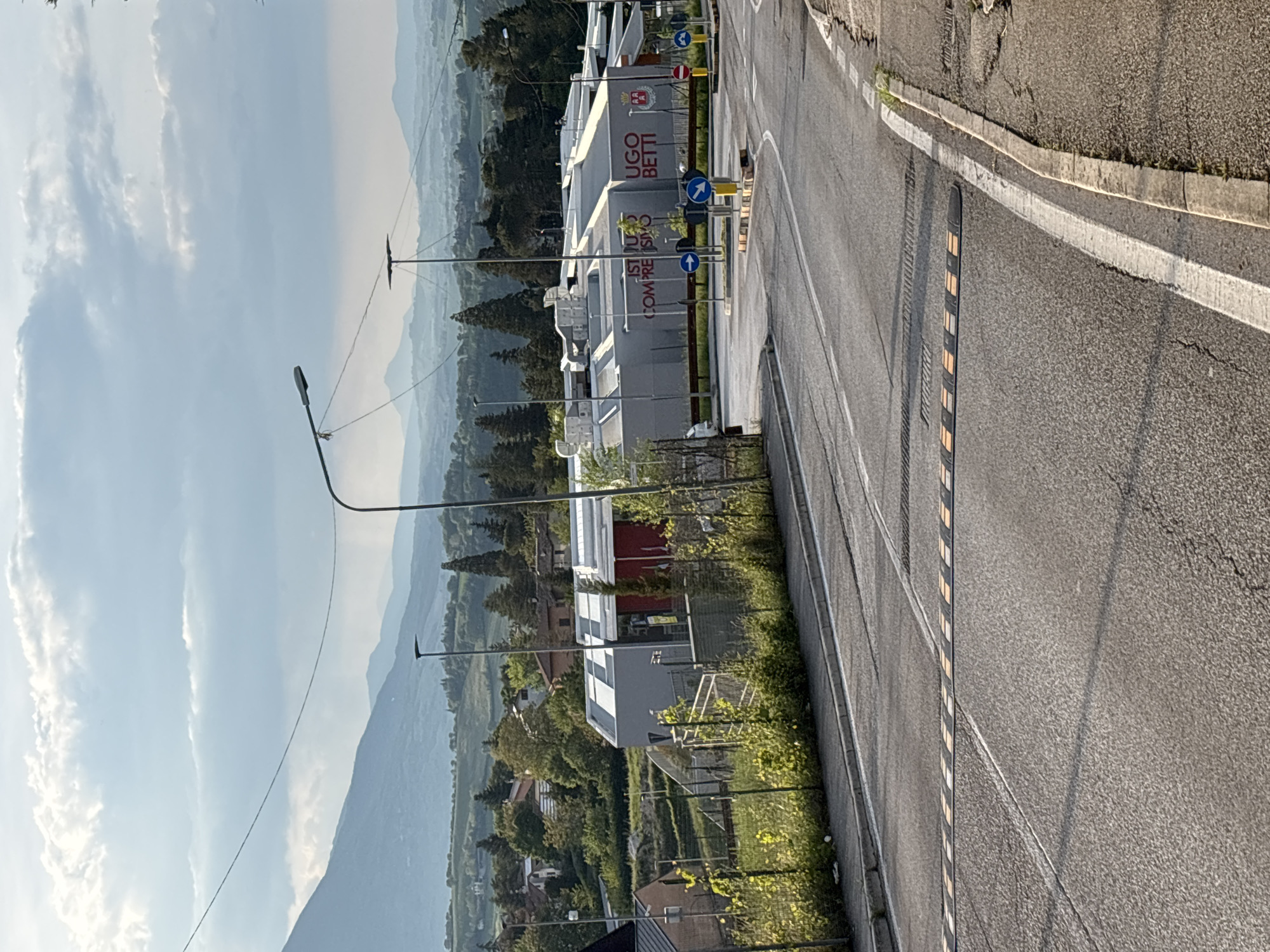 The image, rotated correctly, presents a daytime scene overlooking a valley landscape, likely in a mountainous region such as Camerino, Italy.
In the foreground, a paved asphalt road curves gently, featuring clear white lane markings and a series of yellow and black speed bumps. A concrete curb and grassy shoulder line the right side of the road. Shadows on the road indicate direct sunlight.
The midground is dominated by a modern, light-colored building with red accents. Visible text on its facade clearly reads "ISTITUTO COMPRENSIVO" and "UGO BETTI," suggesting it is a school or educational institution. Several standard European traffic signs are present near the building, including blue circular signs indicating mandatory directions (turn right, straight ahead) and a red circular 'no entry' sign. Surrounding the building are numerous green trees, lush vegetation, and fences. Other buildings with red roofs are visible further back among the trees.
The background features a expansive view of undulating mountains and hills, appearing hazy in the distance. The sky above is partly cloudy with white, wispy clouds against a light blue backdrop. Lampposts and utility lines are scattered throughout the scene. There are no people visible, and no specific event or activity is taking place, depicting a static, clear-weather daytime view.