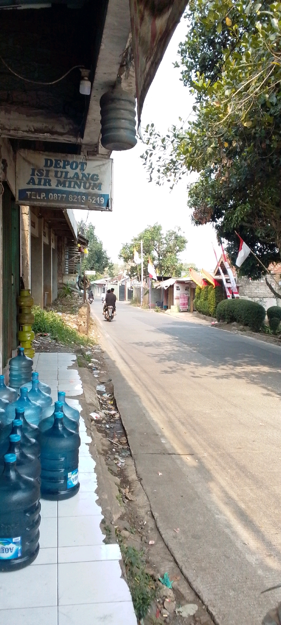 This image depicts a street scene in Cilaku, Indonesia, focusing on a storefront and the adjacent road. The storefront, identified by a sign reading "DEPOT ISI ULANG AIR MINUM" (Refill Drinking Water Depot), displays several large blue water jugs on a tiled sidewalk. To the left of the storefront, a stack of green propane tanks is visible.
The road in front of the depot is paved and shows signs of wear, with some debris along the curb. Trees with lush green foliage line the opposite side of the road, with houses and other small structures visible in the background. Indonesian flags are mounted on poles, suggesting a local celebration or public display.
A motorcycle with two occupants is traveling away from the camera on the right side of the road. The environment appears to be a semi-urban or rural area, characterized by modest buildings, natural vegetation, and a generally well-lit atmosphere indicative of daytime. The overall impression is a snapshot of everyday life in a residential or commercial area of Cilaku.