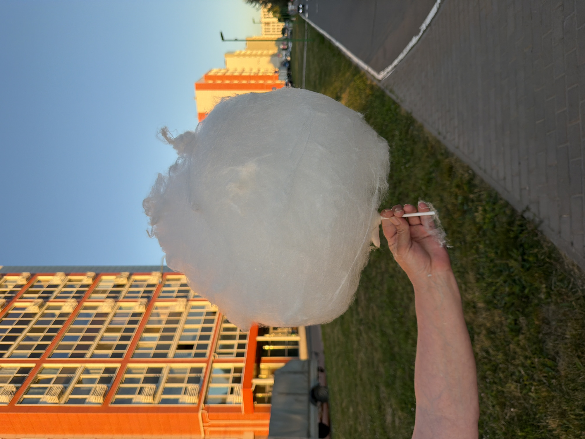 An outdoor, close-up shot focuses on a large, fluffy ball of white cotton candy held by a person's left hand. The cotton candy is the primary subject, dominating the center of the frame and obscuring much of the background. The hand holding the cotton candy is pale, with visible veins. In the background, an orange and white building with many windows is visible, along with a grey truck parked in front of it. To the right of the building, a paved walkway runs alongside a grassy area, with a road and streetlights further in the distance. The scene is illuminated by warm, low-angle sunlight, suggesting it is either sunrise or sunset, casting long shadows and highlighting the textures of the cotton candy and the building. The sky is a clear, pale blue.
