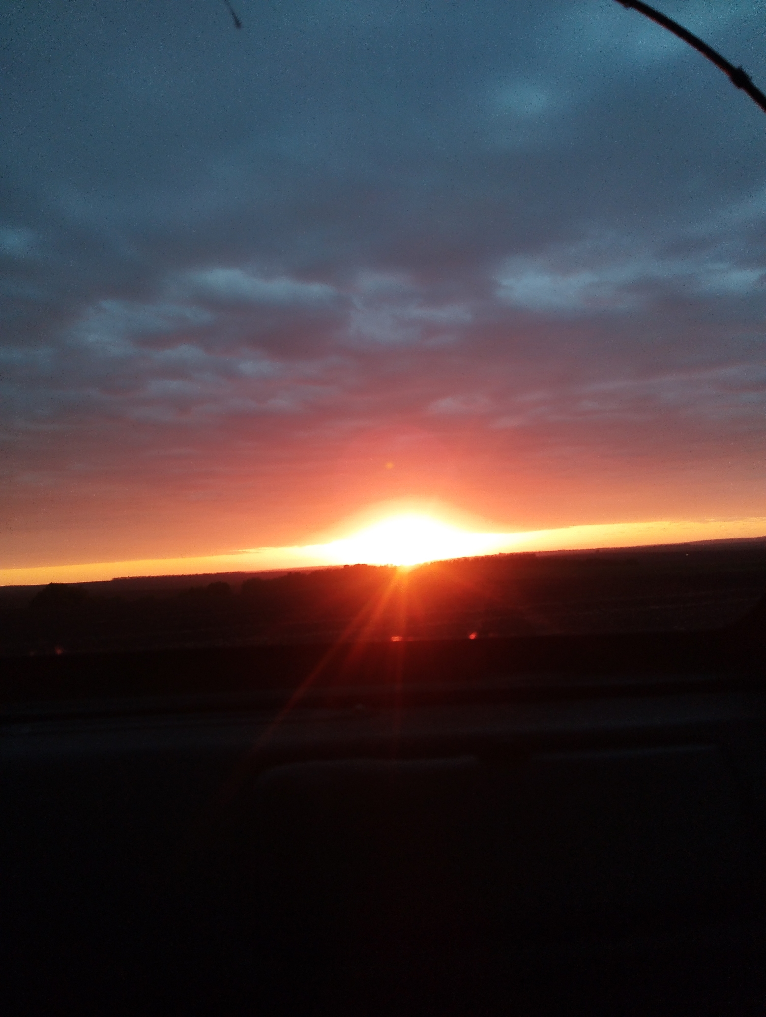 The image shows a vibrant sunset over a dark, silhouetted landscape. The sky is a gradient of dark blue at the top, transitioning to red and orange near the horizon where the sun is setting. A bright beam of light emanates from the sun, casting rays across the landscape. The foreground is dark, possibly representing a road or ground level, and a small branch is visible in the top right corner. The location is Unknown, Unknown.