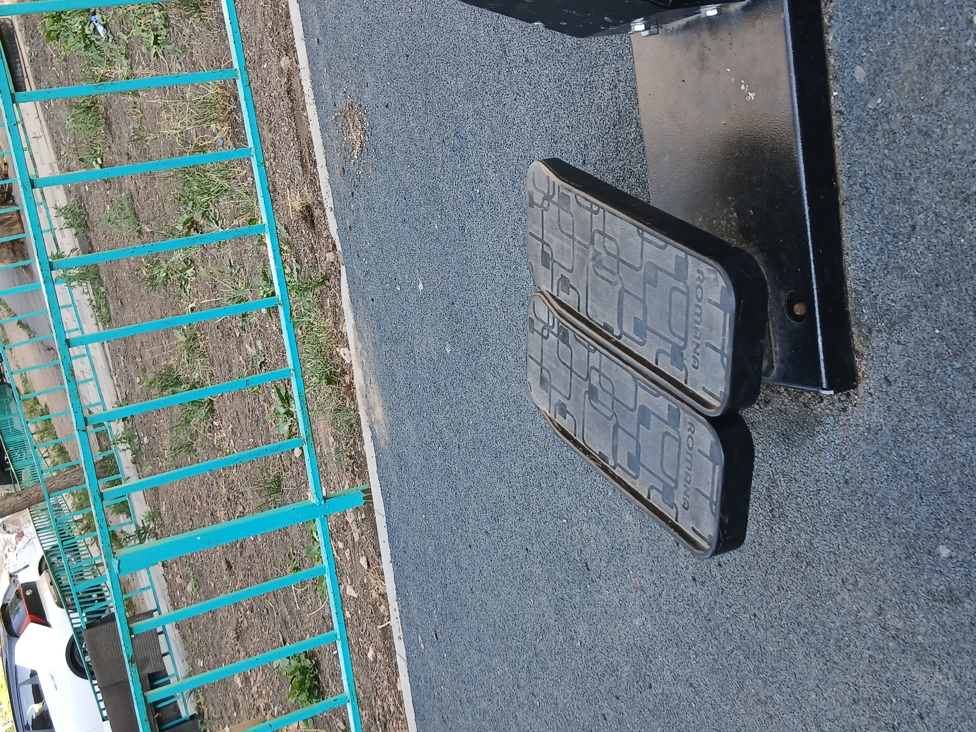 The image shows a close-up view of the pedals of an outdoor elliptical exercise machine. The pedals are black with a patterned surface and the word "ROMANA" embossed on them. They are attached to a black metal frame, which is resting on a dark grey, textured surface, likely a rubberized playground or gym floor. In the background, a turquoise metal fence is visible, with dry grass and dirt visible beyond it. A white car with a black roof and a small black spoiler is partially visible behind the fence, suggesting an urban setting. The overall scene appears to be an outdoor fitness area.
