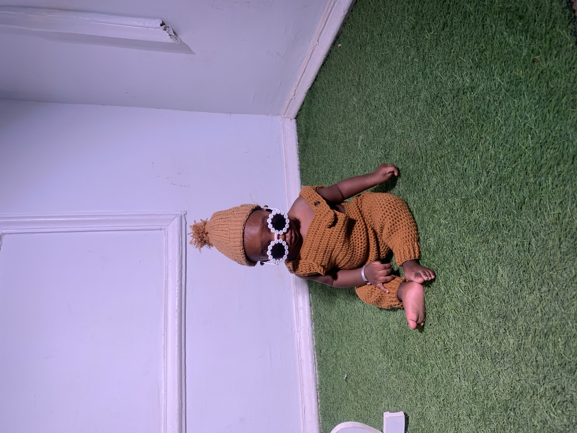 A baby is lying on green artificial turf in a room in Barakin Akawo, Nigeria. The room's walls are white, and the space has a decorative white ceiling fixture. The baby is wearing a brown knitted outfit consisting of a hat with a pom-pom, a romper, and white flower-shaped sunglasses. The baby is resting in the corner of the room, near the baseboard.