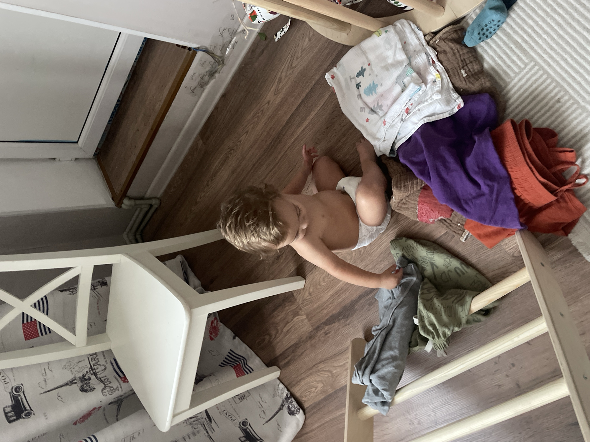 A toddler with blond hair, wearing a diaper, is sitting on a wooden floor next to a white chair. The toddler is reaching for a pile of clothes, including a purple t-shirt, an orange garment, and a green patterned shirt. Several white cloths with a festive pattern are also visible in the pile. A blue sock with grippers is on the floor near the clothes. The room appears to be a child's play area or bedroom, with a wooden climbing structure visible on the right side of the frame. The floor is made of wooden planks. The lighting suggests it is daytime. There is no visible text in the image. The context provided is Almaty, Kazakhstan.