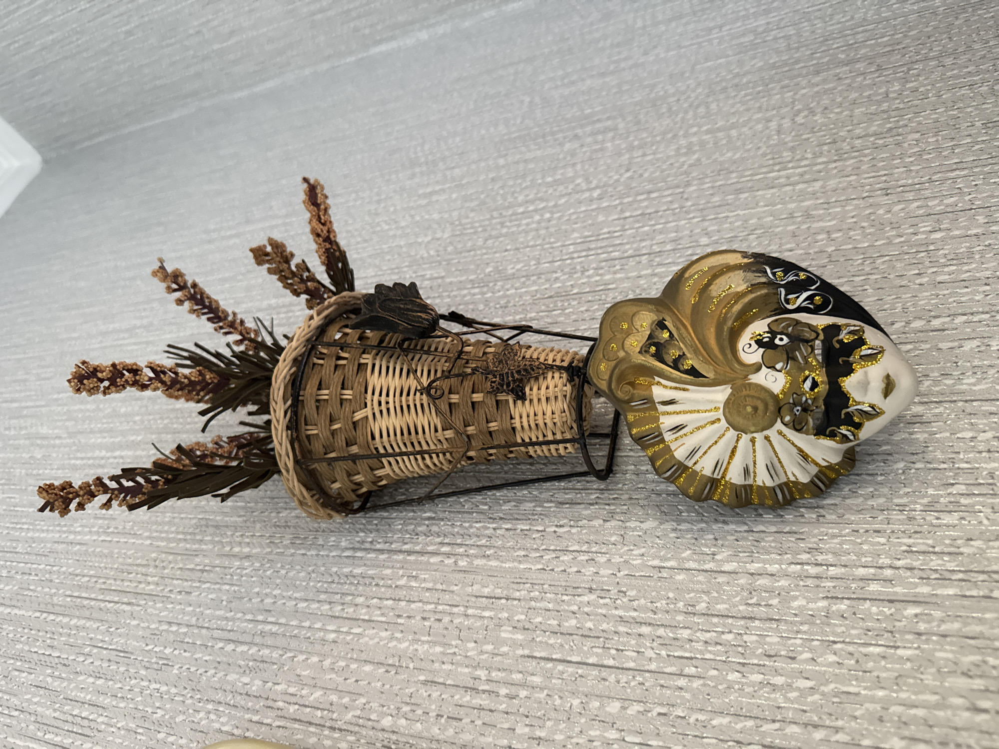 The image displays a decorative wall hanging. The main subject is a small, woven basket mounted on a metal frame attached to a textured, light-colored wall. The basket contains dried, brown floral stems. Below the basket, a Venetian-style mask is suspended. The mask is heart-shaped with intricate detailing in gold, black, and white, featuring a face with painted features and decorative elements resembling a fan or feathers. There are no people or actions depicted in the image, and the environment appears to be an indoor setting, likely a room. The image does not provide any information that would allow for the determination of its location in Shymkent, Kazakhstan.