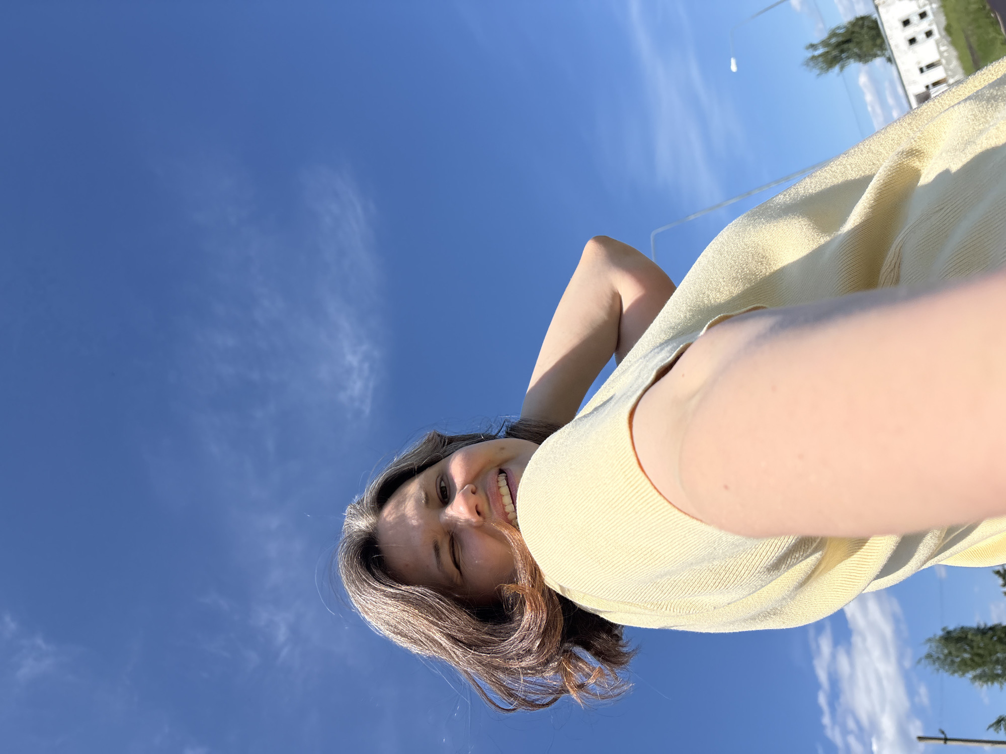 The image displays an adult female subject positioned outdoors in Chaikovsky, Russia. She has shoulder-length, wavy brown hair with distinct gray streaks, and is dressed in a light yellow, ribbed short-sleeved top. The subject is captured looking directly at the camera, winking with her right eye, and smiling with her teeth visible. She is holding the camera in a selfie orientation, with her right arm extended. Her left arm is bent, with the elbow visible near her torso. The scene is brightly lit by natural sunlight, with a predominantly clear, deep blue sky featuring sparse, wispy white clouds. The camera angle is low, looking upward. In the background, on the far left, a dark vertical pole is partially visible. On the right, a white street light pole is present alongside a light-colored building featuring multiple dark window openings, and a tall, slender tree with green foliage, positioned above green ground vegetation.