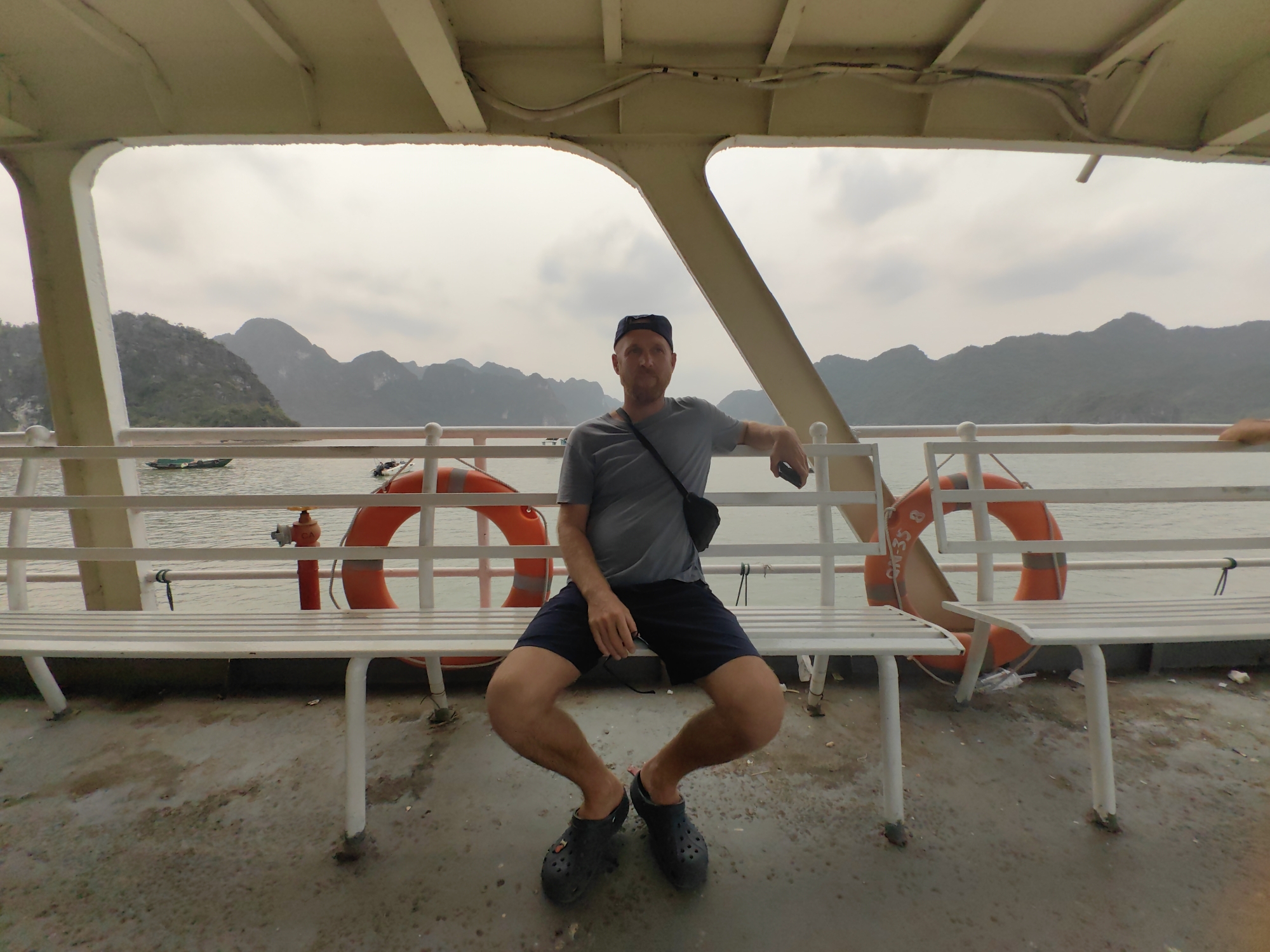 On the deck of a boat, a man sits on a white metal bench, his legs splayed and feet turned inward, shod in dark blue Crocs. He wears a gray t-shirt, dark shorts, and a dark blue baseball cap. A black bag is slung across his chest. In the background, the waters of Hạ Long Bay in Hải Phòng, Vietnam, are visible, dotted with karst formations. The boat's white metal railing and orange life preservers frame the scene. The overhead structure is white, with exposed wiring. The deck floor is stained and appears wet.