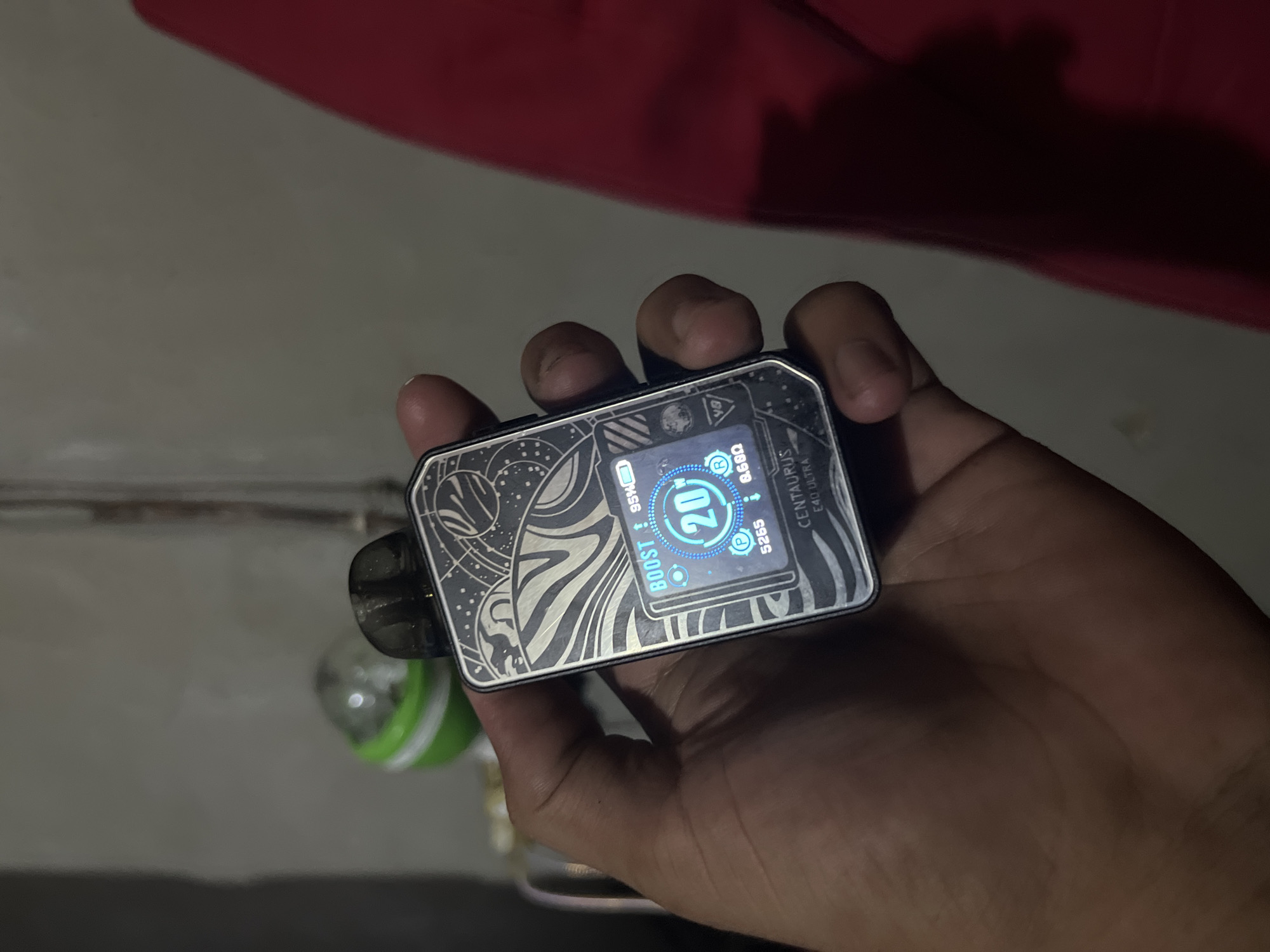 A close-up shot shows a hand holding a vaping device. The vaping device is silver with a black mouthpiece and a colorful digital screen displaying the word "BOOST" at the top left, the number "20" in a large circle in the center, and other icons and text. The background is out of focus and appears to be an indoor setting with a blurred green object on the left and a red fabric on the right. The image does not provide any information that would allow for the determination of its location in Central Jakarta, Indonesia.