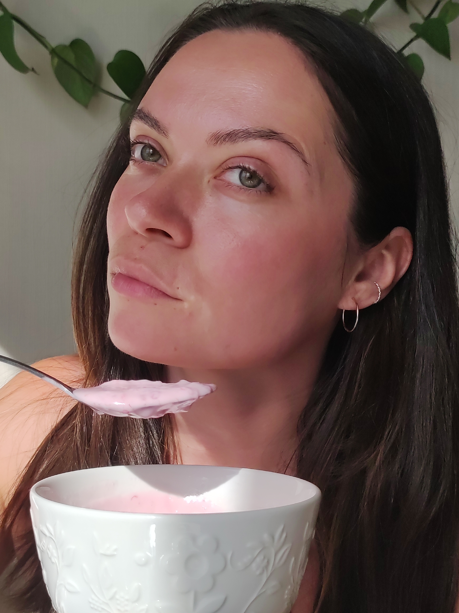 A woman is shown close up in a slightly angled shot, holding a spoon with pink yogurt or smoothie towards her mouth, with a bowl of the same substance below it. She has fair skin, dark hair, and light-colored eyes. She wears a hoop earring in her left ear. The background is blurred, but some green leaves are visible at the top left, suggesting indoor plants. There is no evidence to suggest this photo was taken in Chaikovsky, Russia.