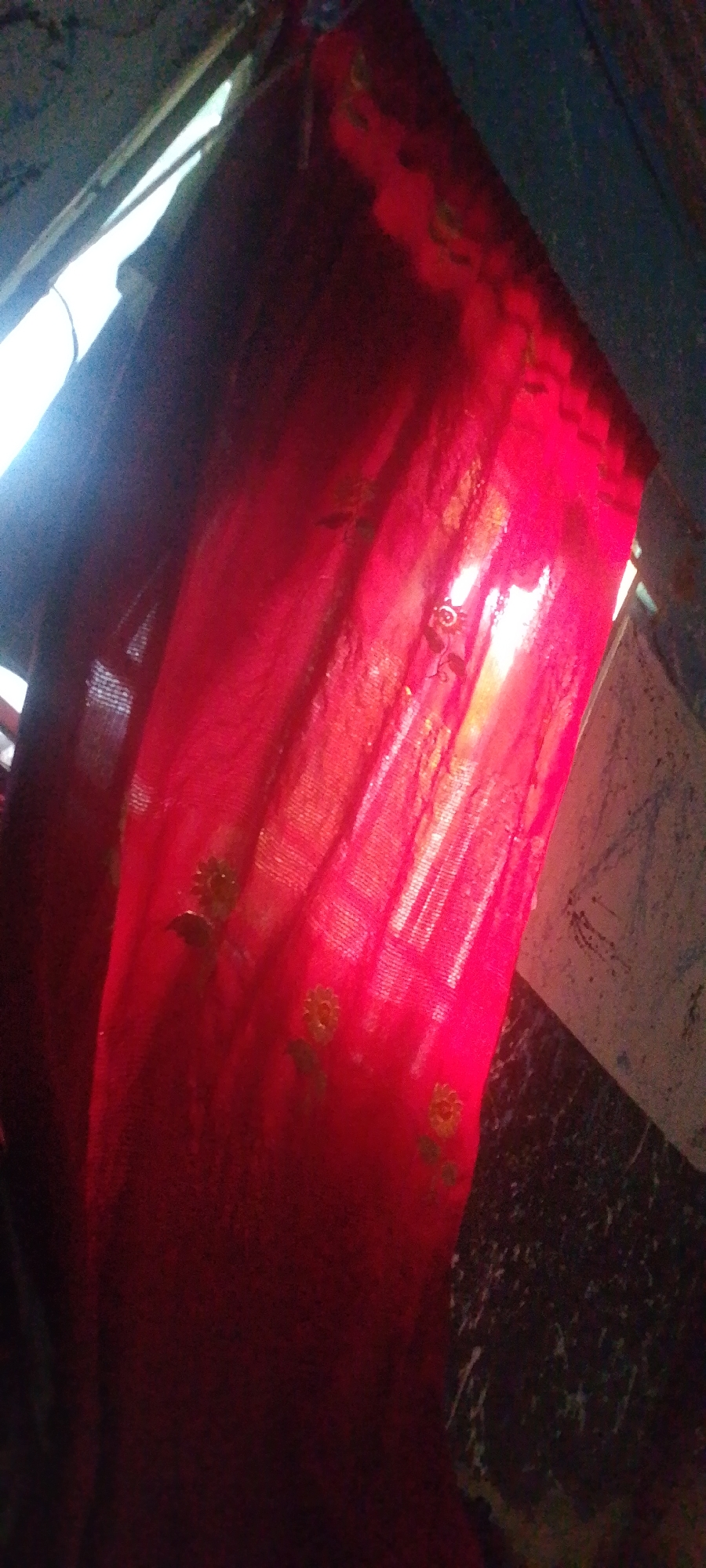 In an indoor environment, a red curtain is illuminated by light from an unseen window. The curtain has a subtle floral pattern, with small yellow flowers and green leaves visible. The fabric appears thin and somewhat sheer, allowing light to filter through, creating bright spots and shadows. The background shows a dark, textured wall. The scene is dimly lit, with the red curtain being the dominant element, suggesting it might be a window covering or a decorative fabric.