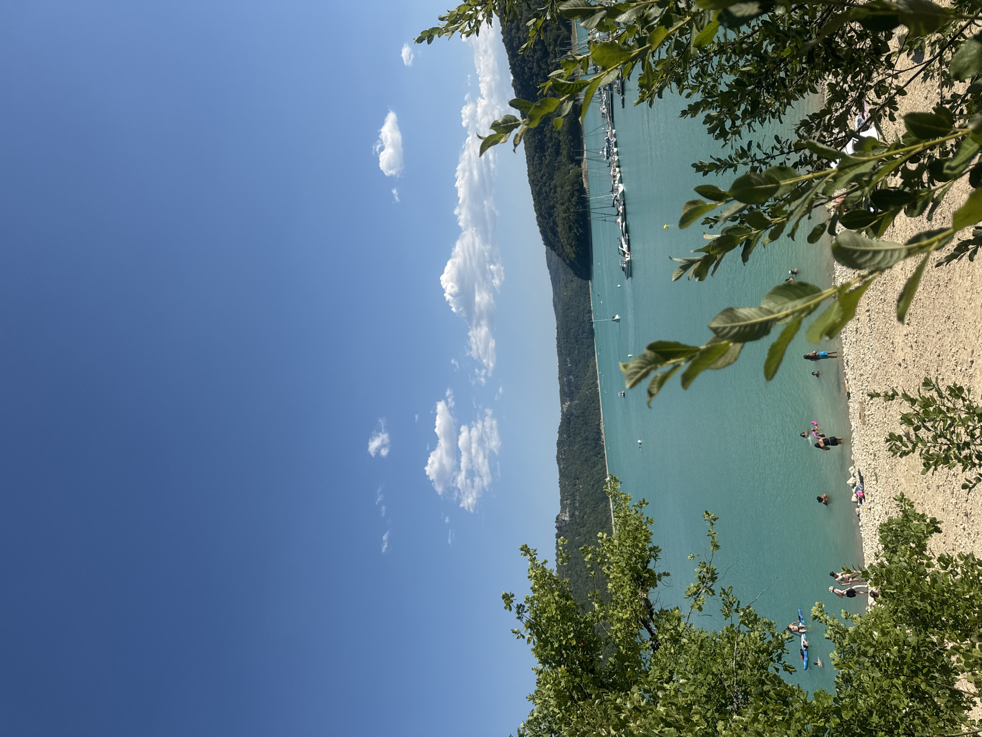This is an eye-level, medium-shot, outdoor photograph taken in Maisod, France, on a clear, sunny day. The scene depicts a body of turquoise water, likely a lake or reservoir, with a shoreline populated by a few people. In the distance, a marina is visible with numerous sailboats and motorboats docked. Lush green hills covered in trees form the backdrop to the water. In the foreground, branches and leaves of trees frame the view, with some leaves in focus and others blurred.
On the beach, several individuals are visible. Some are standing in the shallow water, while others are on the sandy and pebble-strewn shore. One person is lying down on the beach. Further out in the water, individuals are engaged in various activities, including what appears to be paddleboarding. The water is calm and inviting. The sky is a vibrant blue with a few scattered white clouds. The overall impression is one of a peaceful and scenic summer day.
