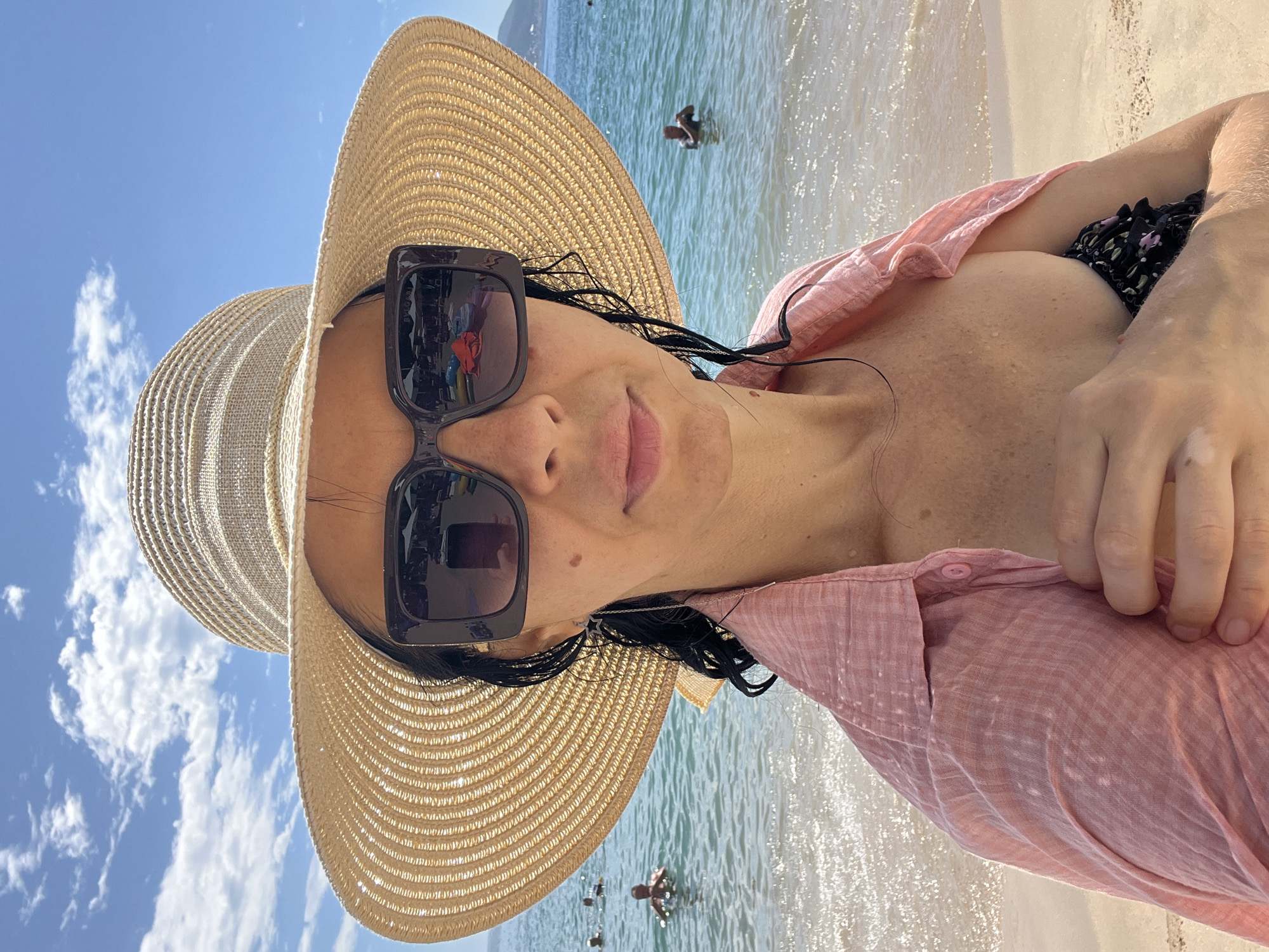 A woman in a large straw hat and sunglasses is taking a selfie on a beach. She is wearing a pink open shirt over a black floral bikini top. Her hair is dark and wet, and she has a star-shaped earring. Behind her, the ocean is clear and blue with gentle waves. Several people are visible in the water in the background. The sky is bright blue with scattered white clouds. The scene suggests a sunny day at the beach in Nha Trang, Vietnam.