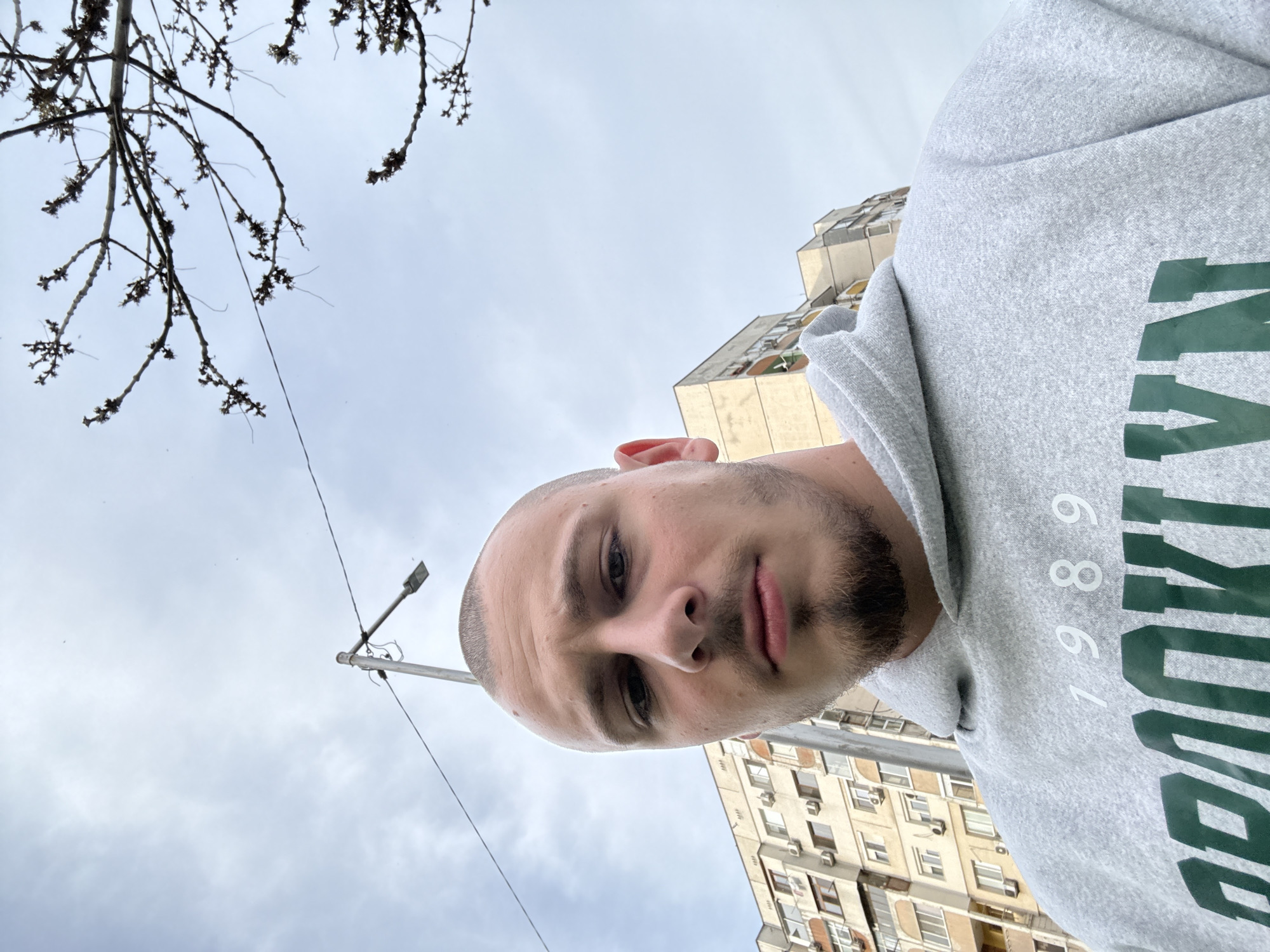 The image is a low-angle, tilted selfie of a young man in an urban setting in Stara Zagora, Bulgaria, taken on an overcast day. The subject is a man with a shaved head and a short beard, appearing to be in his 20s or 30s. He is looking directly at the camera with a neutral expression. He wears a light grey hooded sweatshirt (hoodie) with green text and white numbers printed on the front. When properly oriented, the visible text on the hoodie appears to be "BROOK" or "BROOKLYN" in green letters and the numbers "1989" in white.
In the upper left corner, bare tree branches with small buds are visible against the cloudy sky, suggesting late winter or early spring. A street light pole with a light fixture and a thin wire stretch across the sky in the background, primarily on the left side. The right and lower portions of the background feature several multi-story apartment buildings, characterized by numerous windows, balconies, and visible air conditioning units, typical of residential architecture in the region. The buildings are light-colored. The overall impression is an outdoor selfie taken from an upward-looking perspective in a city environment.