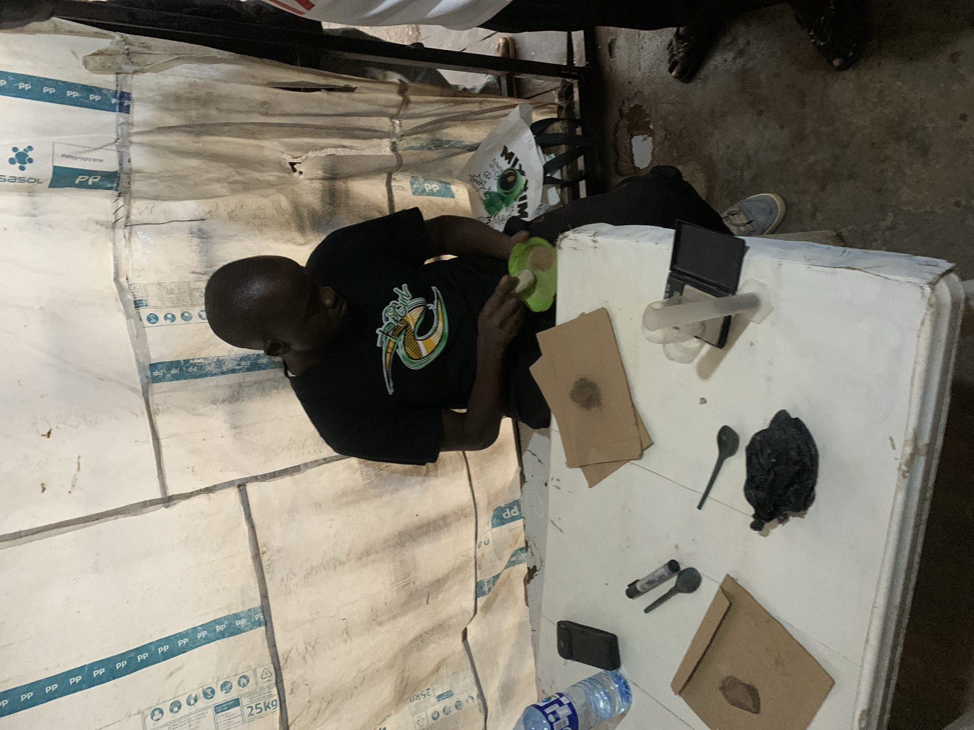 A dark-skinned man wearing a black t-shirt and dark pants sits at a white table in what appears to be a tent or makeshift shelter. He is looking down at a small green bowl in his hands, which contains a light brown powder. A water bottle, a cell phone, and two brown paper envelopes with contents are on the table in front of him. Further to the right, a dark, amorphous object is on the table, along with two black spoons and a marker. The background consists of a white tarp-like material with blue markings and text, and the floor is a rough, dirty concrete surface with some puddles. The overall scene suggests a clandestine operation or processing of materials.