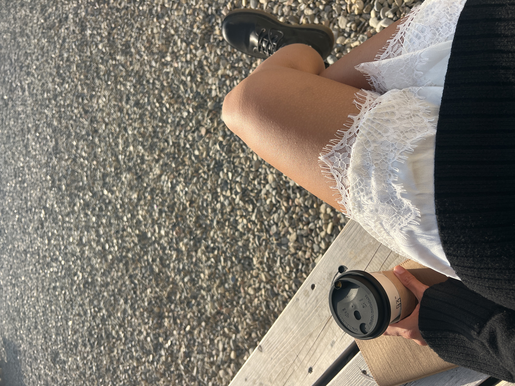 The image shows a person sitting on a wooden bench in what appears to be an outdoor setting. The person is wearing a white dress with lace trim and a black sweater, and has a black shoe visible on one foot. They are holding a takeaway coffee cup in their hand. The ground surrounding the bench appears to be covered with small pebbles or gravel. The location is not identified.