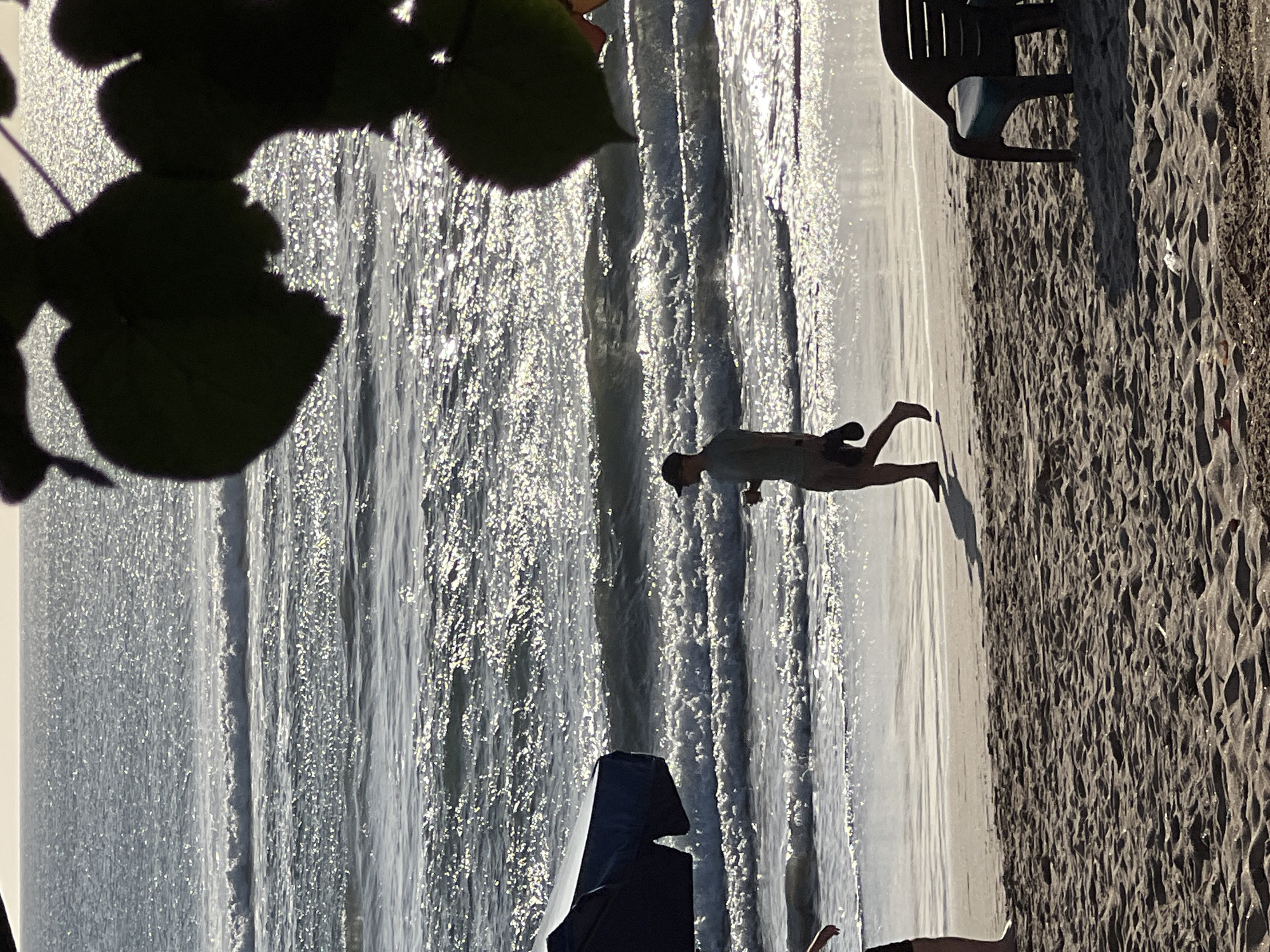 A lone figure walks along the shoreline in Kuta, Indonesia, as waves crash onto the sandy beach. The sun, high in the sky, casts a shimmering reflection on the ocean's surface. In the foreground, a black plastic beach chair sits empty, awaiting a visitor. The sand is textured with ripples, hinting at the ebb and flow of the tide. In the background, lush green foliage is silhouetted against the bright sky, framing the scene. The overall impression is one of tranquility and natural beauty.