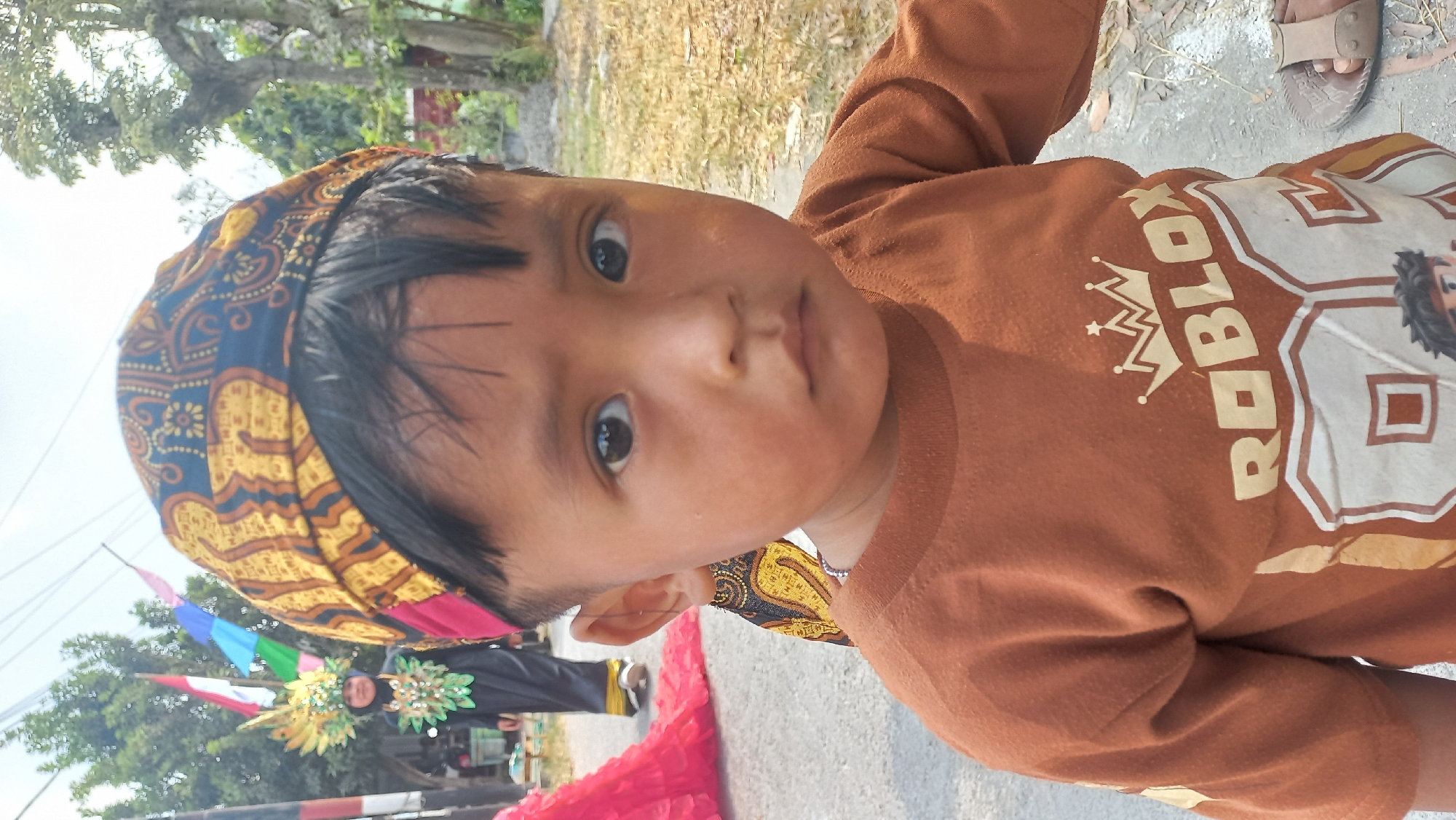 A close-up, eye-level outdoor portrait captures a young boy in the foreground, facing the camera with a neutral expression. He has dark hair and dark eyes, and is wearing a brown long-sleeved t-shirt with a graphic that appears to say "ROBLOX" above the number "89" and an image of a cartoon character. He is also wearing a traditional patterned head covering with yellow, black, and red elements.
In the background, slightly out of focus, a person in traditional attire, possibly a dancer, is visible on the left. This person is wearing a black outfit adorned with green and gold embellishments and a tall, elaborate headdress with golden feathers and colorful flags. The setting appears to be a street or pathway, with dry grass, trees, and foliage visible. The overall atmosphere suggests an outdoor event or celebration, possibly in a rural or semi-urban area like Kedungmegarih, Indonesia, given the cultural attire and the presence of flags. The lighting indicates it is daytime, likely with bright sunlight.