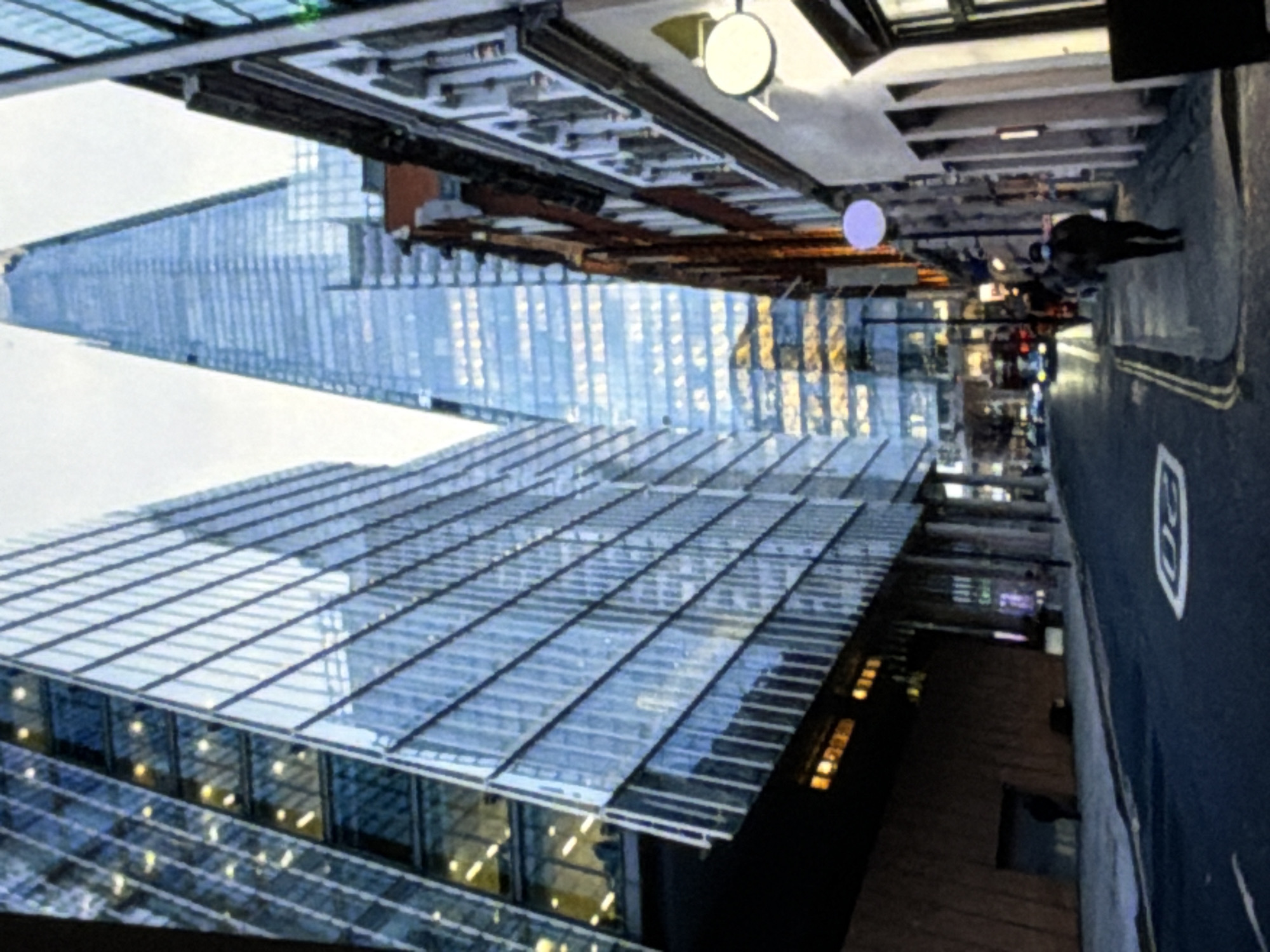 The image displays a street scene within an urban environment. On the left, a multi-story building with a facade of reflective, blue-tinted glass stands prominently, showing illuminated interior spaces. In the background, the distinctly tapered, glass-clad skyscraper known as The Shard is visible, with warm lights illuminating its lower levels. To the right, a row of older, multi-story buildings, featuring traditional brick and stone construction with multiple windows, lines a sidewalk.
A paved street runs through the foreground and middle ground, displaying a white "20" speed limit road marking and double yellow lines along the left curb. Further down the street, multiple vehicles are visible. On the right-hand sidewalk, a lone individual in dark clothing and a blue face mask walks away from the viewer. A second, less distinct figure in dark attire is partially visible near the base of the glass building on the left. A round, light-colored sign is affixed to a building on the right. The lighting indicates an overcast day or early evening. This scene is located in London, United Kingdom, as identified by the presence of The Shard.