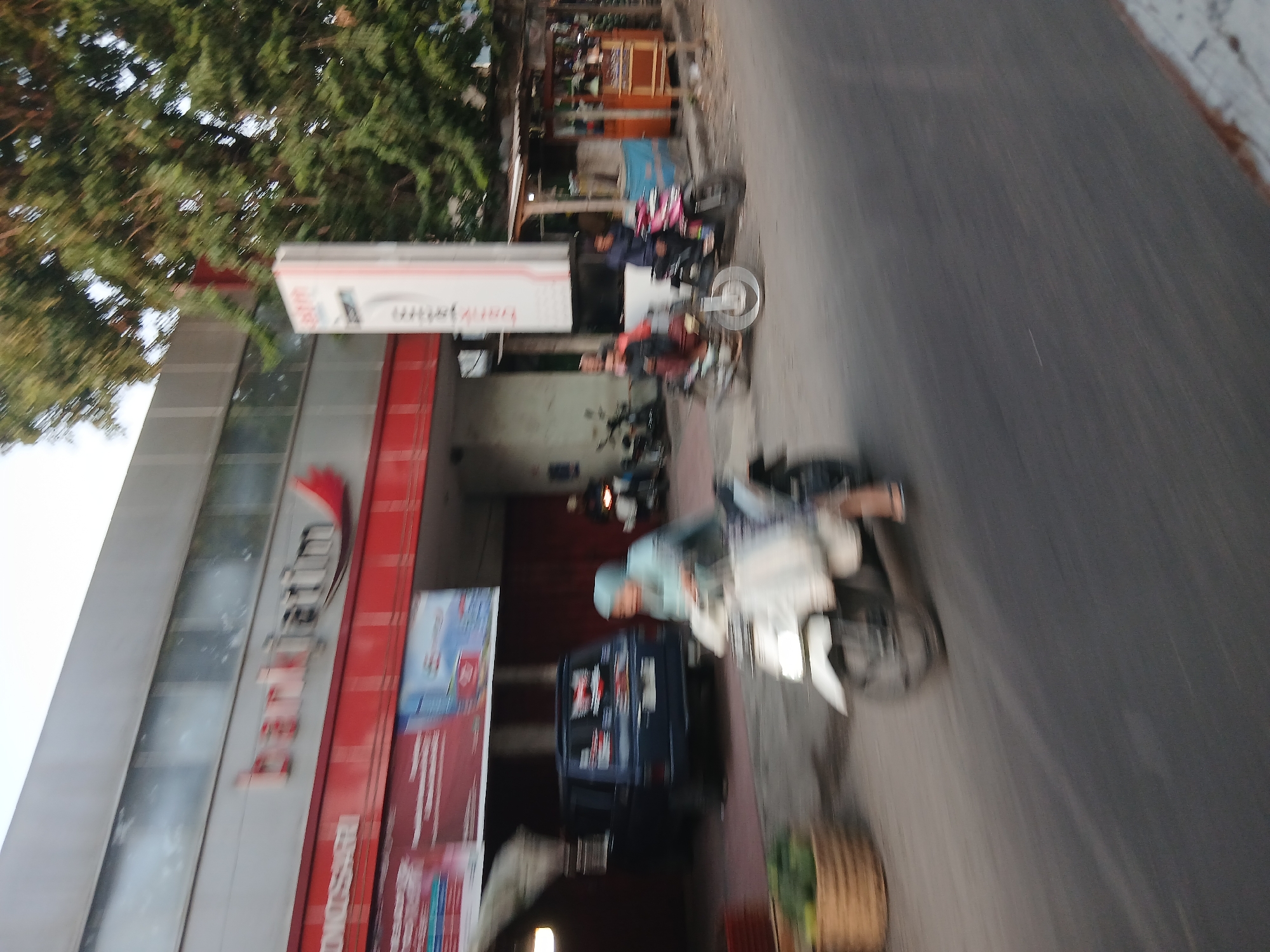 The image captures a street scene in Wonosari, Indonesia, with a focus on a bank building and passing traffic. In the foreground, a person on a white scooter is moving from left to right, their body and the scooter blurred due to motion. To the left of the scooter, a dark blue van is parked. Behind the van, the facade of a building is visible, featuring a large red and white sign with the text "bankjatim" in red letters. The building has a grey, horizontal paneling and windows. In the middle ground, several other motorbikes are present, with some people on them, also showing signs of motion blur. One motorbike is pink and black, and another is a lighter color. To the right, a small wooden stall with a blue tarp overhead is visible, with items displayed on shelves. Lush green trees with dense foliage are present behind the stall and to the right of the bank building. The ground is paved with asphalt. The lighting suggests it is daytime, possibly late afternoon or early morning, with soft, diffused light.