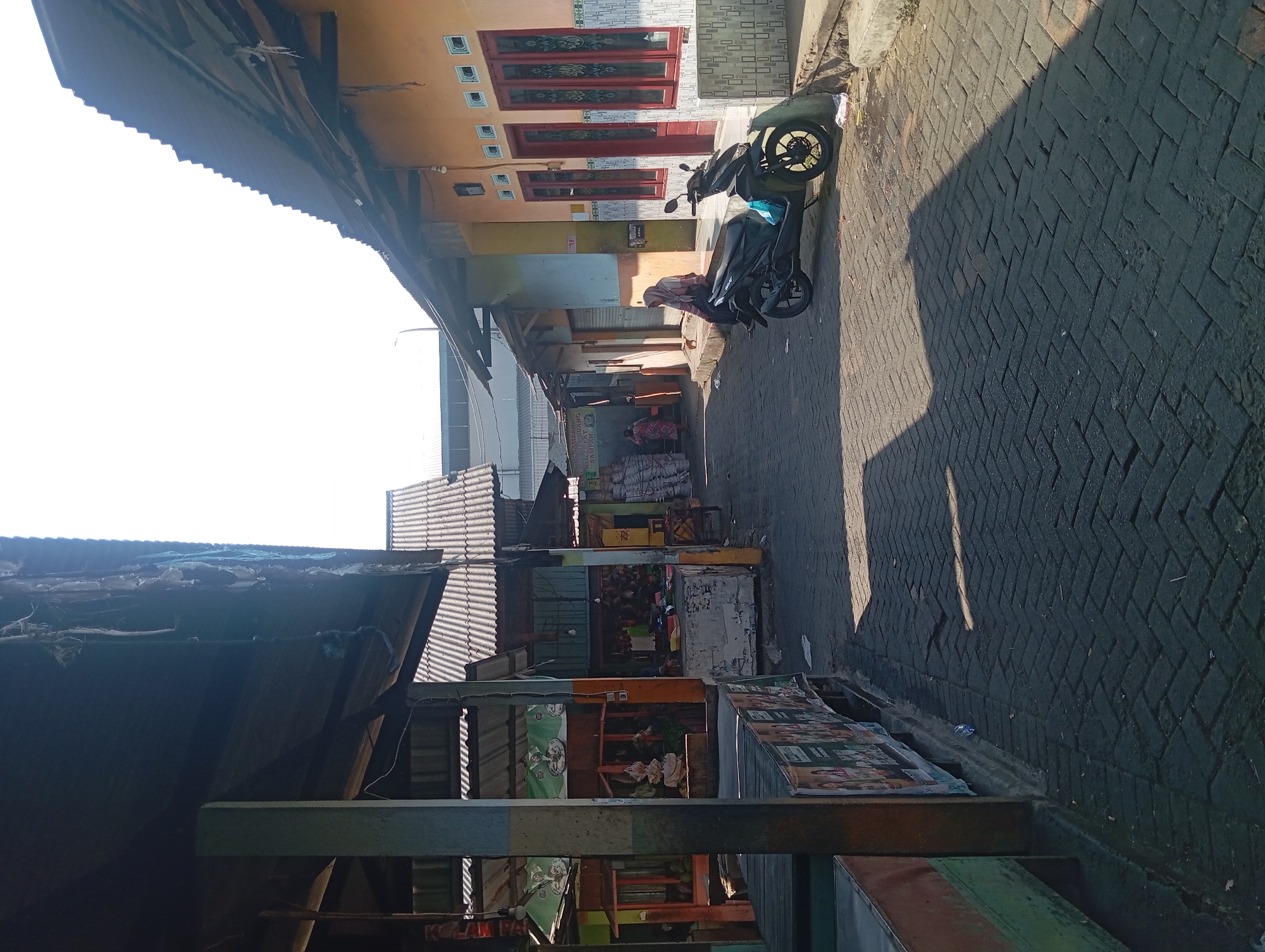 The image depicts a street scene in Kepulungan, Indonesia, likely a market area. On the left, a weathered wooden stall with a corrugated metal roof is visible, exhibiting peeling paint and some disarray. Posters are affixed to its front. Further down the street, more stalls with similar roofing are present, displaying various goods and some raw produce. A motorcycle, black and parked on the right side of the street, is prominent in the mid-ground. A woman wearing a hijab sits on the curb next to the motorcycle. The street is paved with interlocking dark bricks arranged in a herringbone pattern, with strong shadows cast from the structures and the motorcycle, indicating a bright, sunny day. The buildings lining the right side of the street are painted in warm, earthy tones with decorative tiled elements around the windows and doors. The sky is bright and mostly clear.