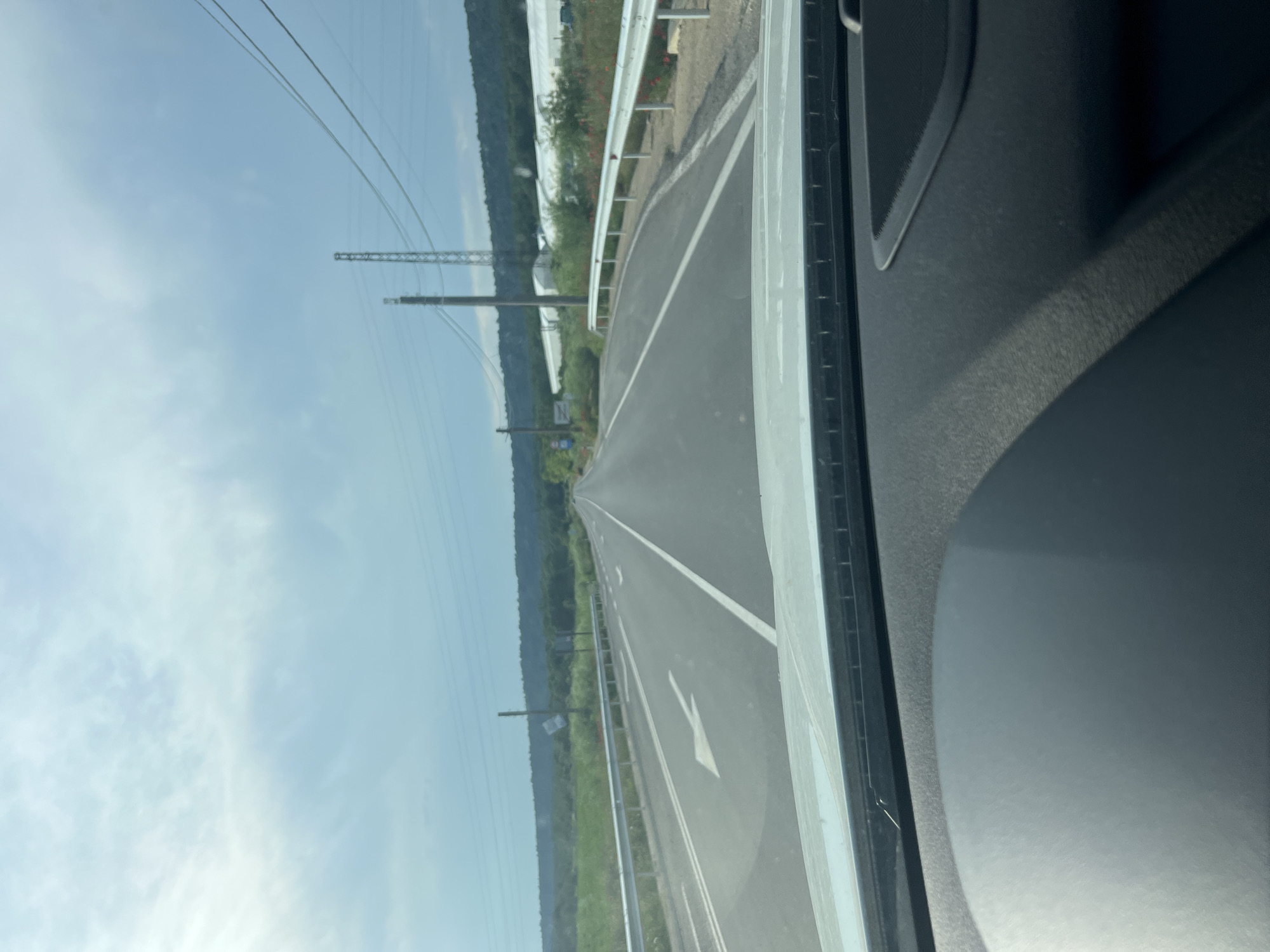 The image captures a forward-facing perspective from inside a vehicle on an asphalt road in the vicinity of Tsarkva, Bulgaria, under a partly cloudy sky. The road is a two-lane thoroughfare with white painted markings: a solid white line delineating the right shoulder, and a double white center line consisting of a solid line on the right and a dashed line on the left, indicating passing is permitted in the direction of travel. A large white arrow on the pavement in the left lane indicates the direction of travel is straight ahead.
To the left of the road, a silver metal guardrail runs parallel to the pavement. Beyond the guardrail, dense green vegetation borders the road, with a blue road sign mounted on a wooden utility pole, partially obscured and indistinct. Further down the road on the left, another white sign is visible on a pole. In the background on the left, a range of hills covered with dark green foliage extends horizontally.
To the right of the road, an identical silver metal guardrail is present, with patches of red flowers (likely poppies) growing along the embankment. Beyond the guardrail and vegetation, a series of large, white, tunnel-shaped agricultural structures, identifiable as greenhouses, are clearly visible, stretching into the distance. In the sky above the right side, multiple power lines are strung, supported by various utility poles, including a tall metal lattice tower and several wooden poles.
The sky occupies the upper portion of the frame, displaying scattered white clouds against a pale blue background. The lower portion of the image reveals the interior of the vehicle, specifically the dark gray dashboard with a textured surface and the lower edge of the windshield. No individuals or specific actions are discernible within the frame.