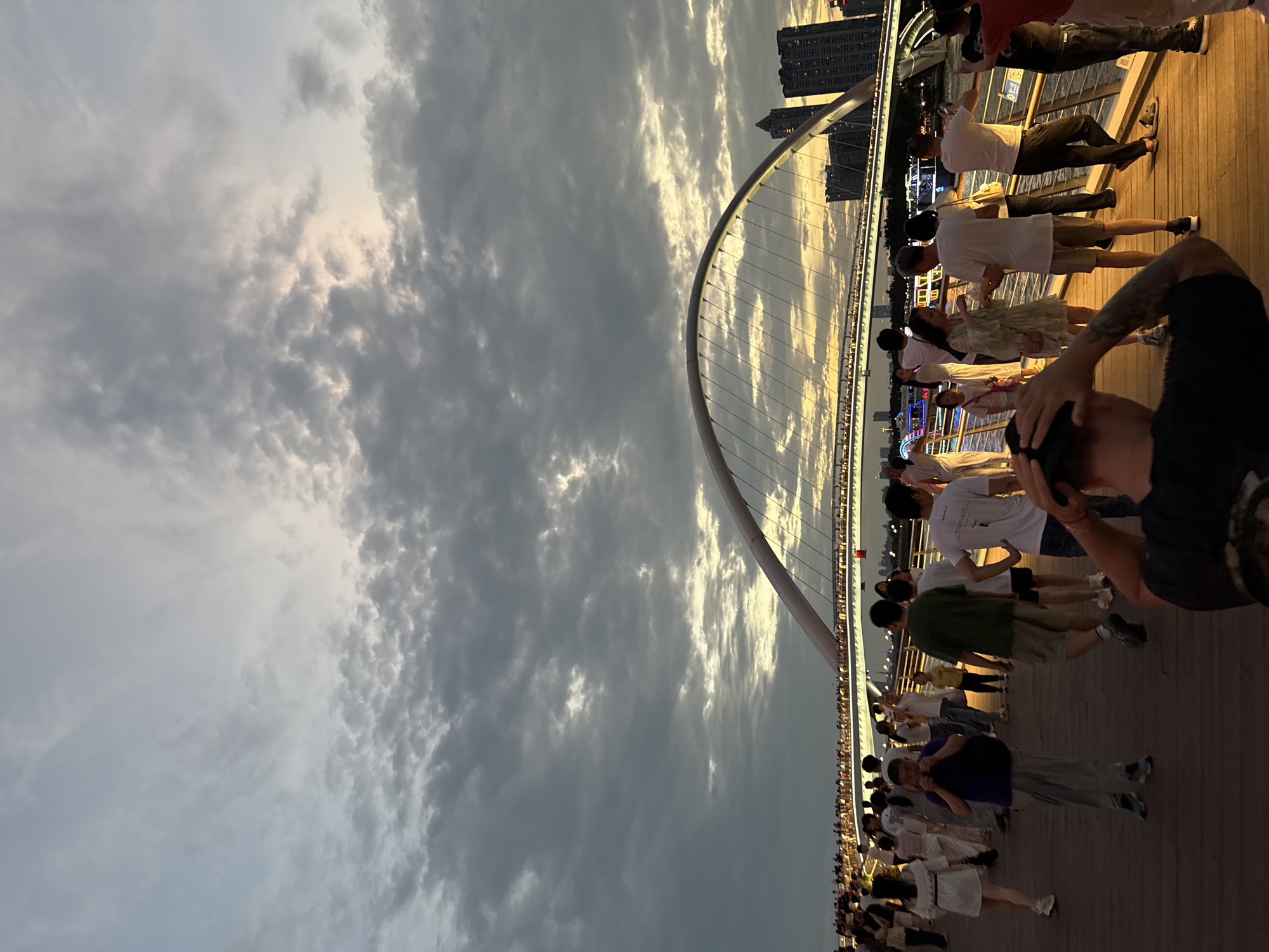 The image shows a crowd of people on a bridge, likely the Liede Bridge in Yuexiu District, China, judging by the arched structure and the city skyline. People are standing and walking, some taking photos. The sky is cloudy.
