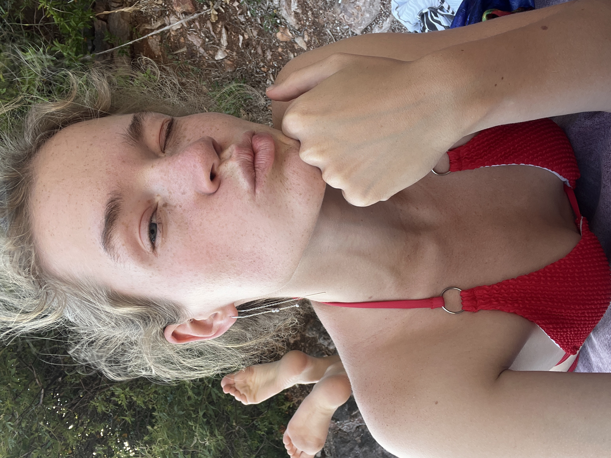 A close-up, eye-level shot depicts an adult female positioned outdoors in Rađenovići, Montenegro. The woman, exhibiting fair skin and prominent freckles across her face, neck, and shoulders, is seen winking her right eye and puckering her lips in a playful expression. Her left hand is resting under her chin, with her fingers curled. Her light-colored, curly hair is styled naturally. She wears a textured red bikini top, featuring a silver O-ring connector on the strap visible over her right shoulder. A long, threader-style silver earring with small metallic beads or coils is present in her left ear. Her bare feet are visible in the upper left background, suggesting she is lying prone or semi-prone. The background to her left features dense green foliage, while the ground to her right consists of dry earth, small rocks, and sparse vegetation. A purple and blue striped towel or fabric is visible beneath her lower right side. The scene is illuminated by natural daylight.