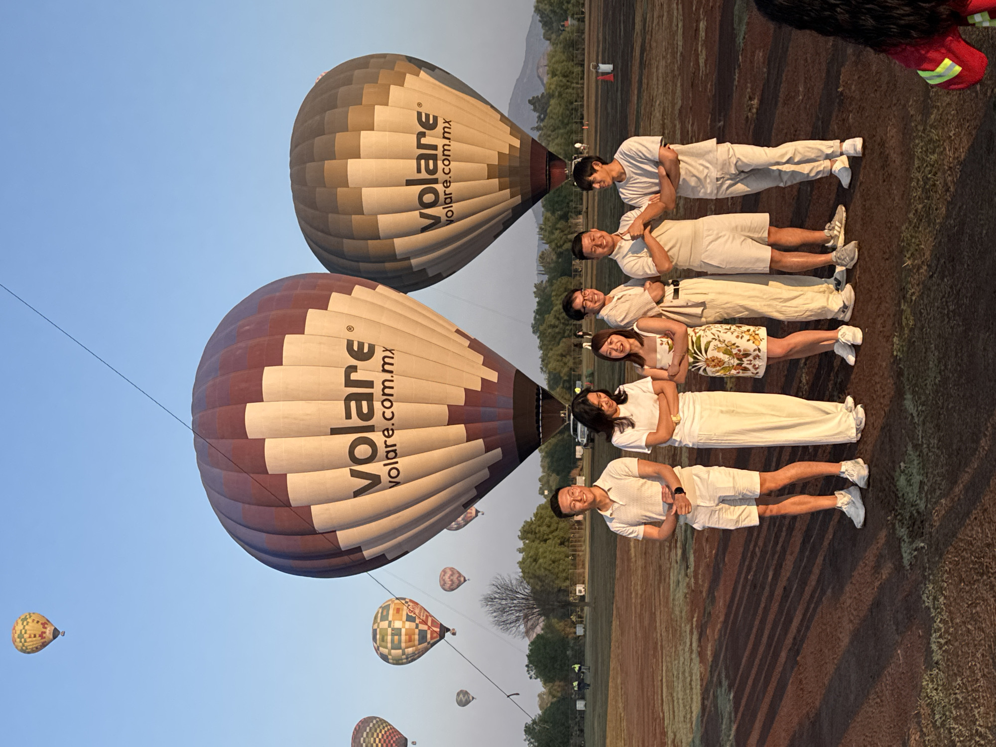 The image depicts a group of six people posing in front of two hot air balloons. The balloons, located in Ampliación San Francisco, Mexico, are labeled "volare.com.mx." The people are wearing casual clothing, mostly in light colors, and are lined up horizontally. Several other hot air balloons are visible in the background, floating in a clear sky. The ground appears to be a field with some sparse vegetation.