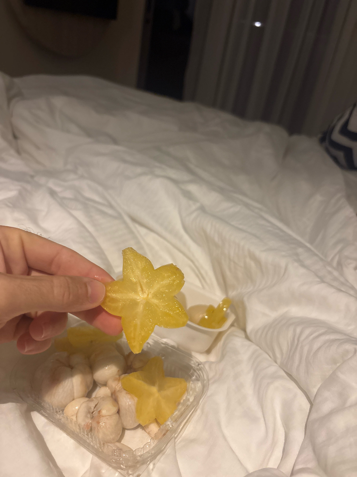 A close-up, waist-level shot shows a person's hand holding a slice of starfruit over a plastic container filled with more starfruit slices and what appear to be mangosteens. The items are arranged on a rumpled white duvet, suggesting a bed. The background is dimly lit with curtains visible in the upper right corner, hinting at a hotel room or bedroom. The scene is devoid of any identifiable urban elements of Karon, Thailand, as it takes place entirely indoors on a bed.
