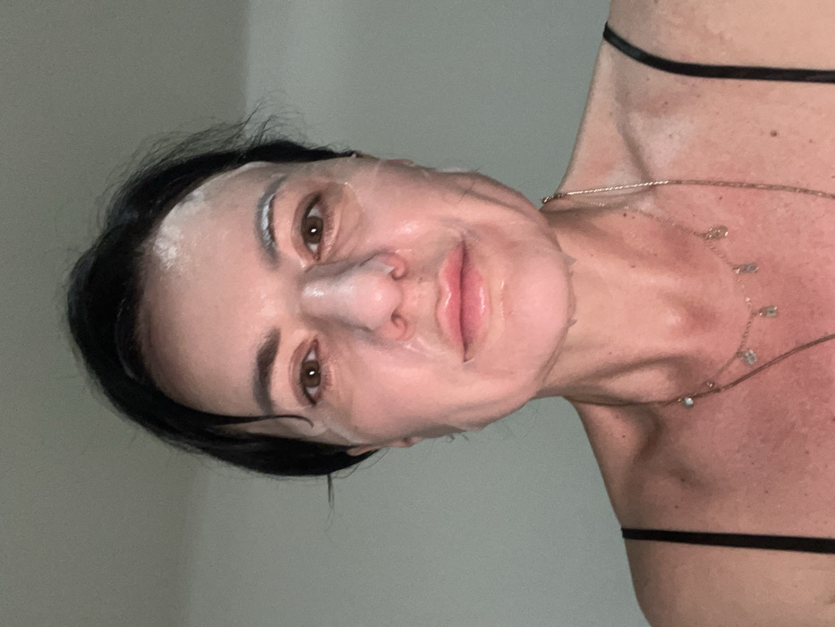 The image displays a close-up, frontal portrait of an adult female situated in a plain indoor environment. Her face is entirely covered by a translucent sheet mask, which is saturated with a liquid substance, leaving apertures for her eyes, nose, and mouth. Her eyes are brown, and her dark hair is pulled back from her face. She is wearing a black garment with thin straps visible over her shoulders. Around her neck, two gold-colored chain necklaces are present: one features multiple small, light-colored charms, and the other is a longer, unadorned chain. The background is a uniform, light grey wall, providing no specific identifying features of a room or structure. The precise location within Smederevska Palanka Municipality, Serbia, cannot be determined from the visual information presented.