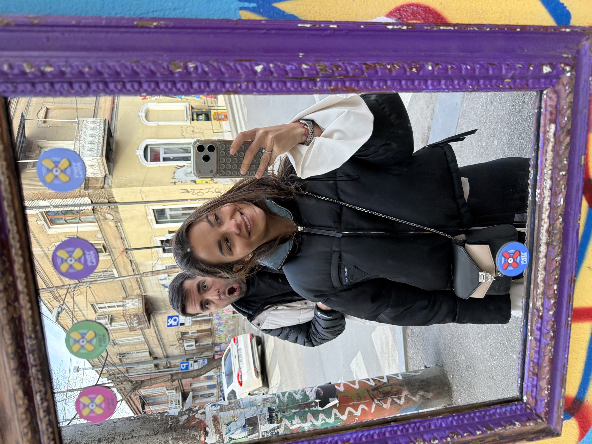 The image shows a reflection in a purple-framed mirror of a woman taking a selfie with a man standing behind her, with an open mouth. The background of the reflection reveals a street scene in Tbilisi, Georgia. Buildings are visible across the street. Street signs are visible, and parked cars line the street. The mirror's frame is ornate and shows signs of wear. Stickers with yellow blades on a background are placed on a pole. The woman wears a black puffer jacket and white blouse, and a small purse. The man is behind and to her left. The selfie is being taken with a smartphone. The mirror is mounted on a decorated surface with colorful graffiti-like designs.
