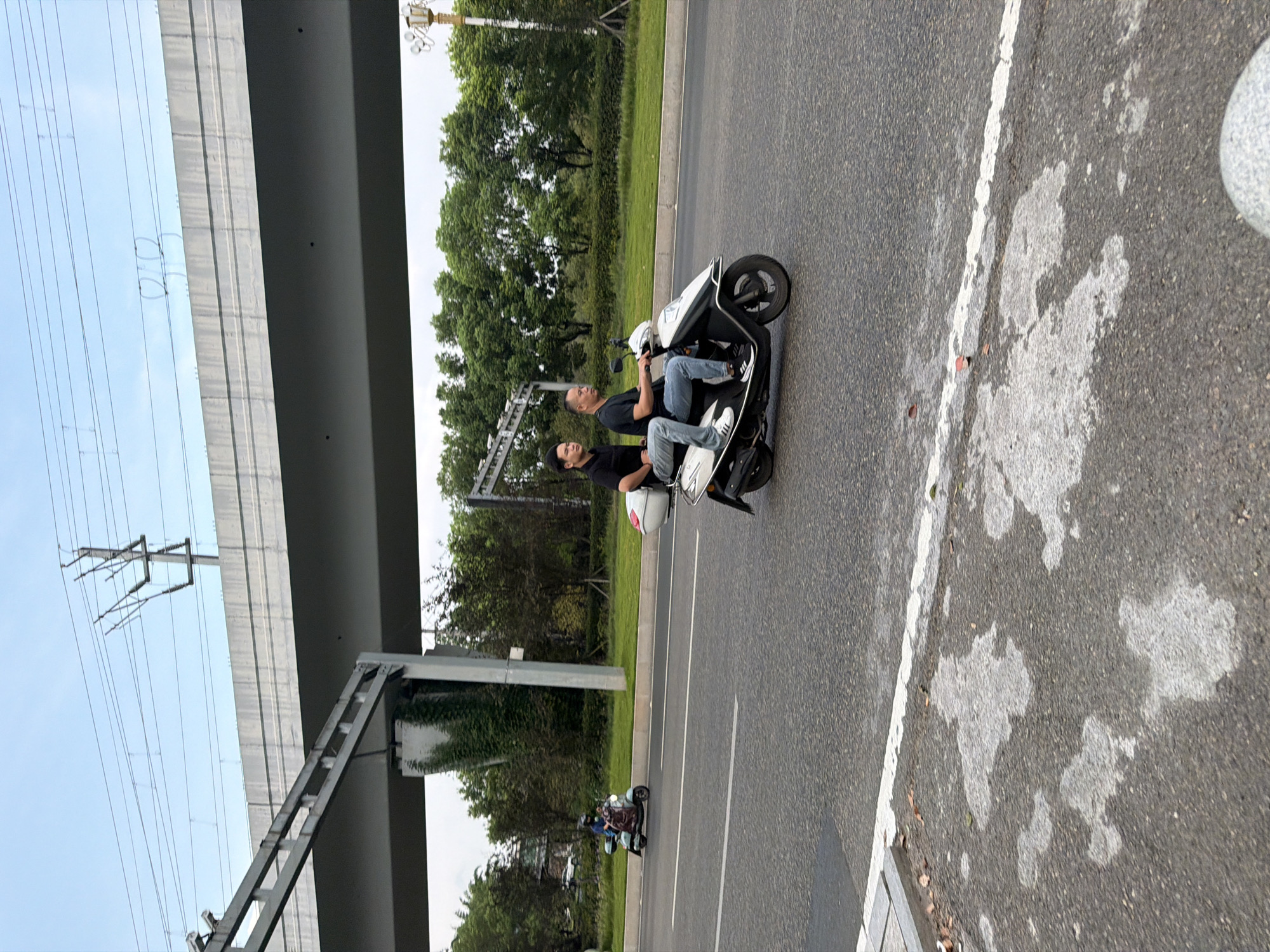 The image depicts a street scene in Jiangjin, China, featuring two individuals riding a scooter on a paved road. The scooter is white and black, with a white storage box on the back. The driver, an adult male, is wearing a black t-shirt and blue jeans. The passenger, a younger male, is wearing a black t-shirt and blue jeans. They are traveling from right to left in the frame.
In the background, a multi-level overpass dominates the scene, with power lines and utility poles visible above it. The overpass is constructed of concrete and metal. Beyond the overpass, lush green trees form a dense line. Another scooter, a lighter color, is visible in the far left of the image, carrying two individuals. The road surface is asphalt, with a white line marking the lane. Some patches of water or discoloration are visible on the road in the foreground. The sky is overcast with a light blue hue.