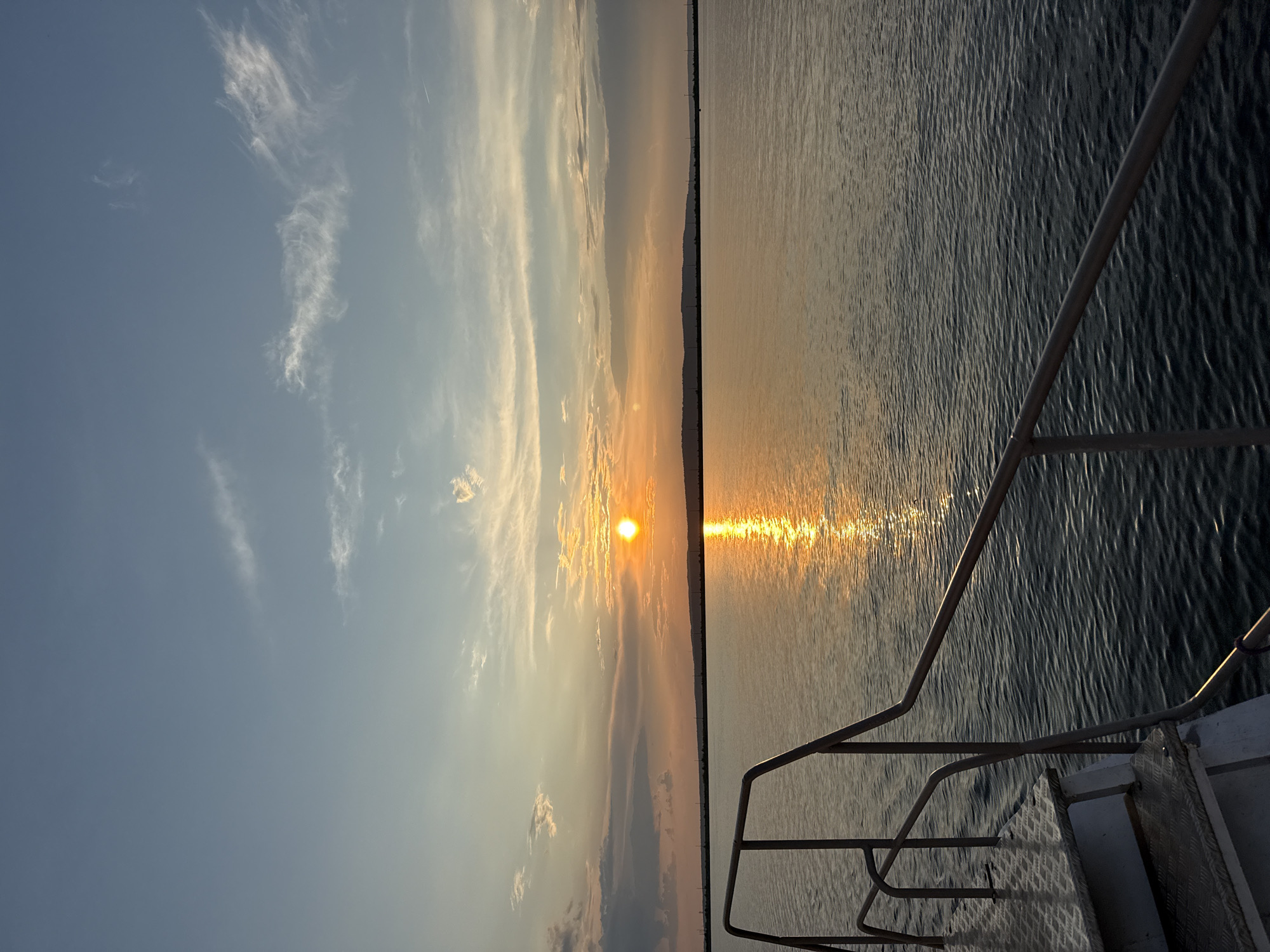 The image depicts a sunset over a body of water, likely a reservoir or river, viewed from the deck of a boat. The sun, a bright orange orb, is low on the horizon, casting a golden reflection on the rippling water. The sky is a gradient of blue and orange, with scattered wispy clouds. In the foreground, a portion of the boat's railing and steps are visible, suggesting the perspective is from a vessel. The distant shoreline is lined with faint silhouettes of trees and possibly structures, under a hazy sky. The scene appears tranquil and serene, bathed in the warm light of the setting sun. This view is consistent with locations along the Kama River in Chaikovsky, Russia, known for its expansive water bodies and scenic sunsets.