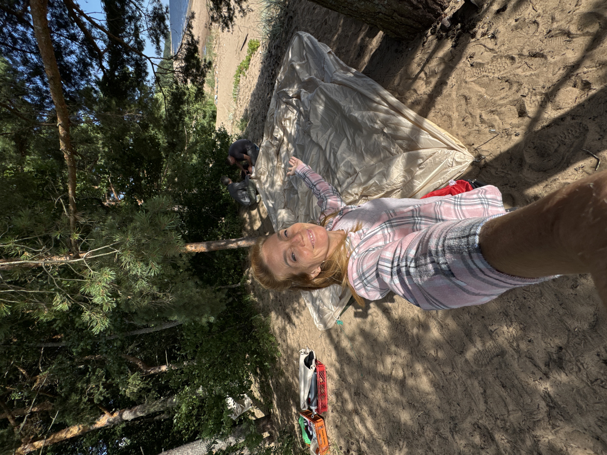 A woman with blonde hair, wearing a pink and white plaid shirt, takes a selfie on a sandy beach in Zelenaya Roshcha, Russia. She is smiling and gesturing with her left hand. In the background, a large, tan tent is spread out on the sand. Two men are crouched near the tent, appearing to be setting it up. Several red crates filled with items are placed to the left of the woman. The environment is a beach with trees and the sea visible in the distance under a clear blue sky.