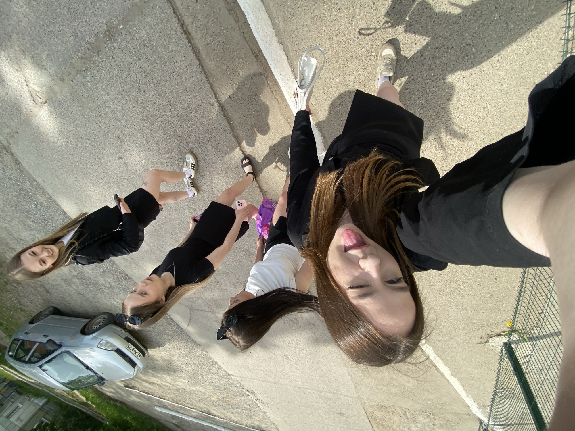 Four young women are in a selfie on an urban sidewalk in Chaikovsky, Russia. They are near a silver car parked on the street, and a fence and grass patch are visible in the background. All the women are wearing variations of black clothing; some have on white shirts or blouses. One is sticking her tongue out and winking.