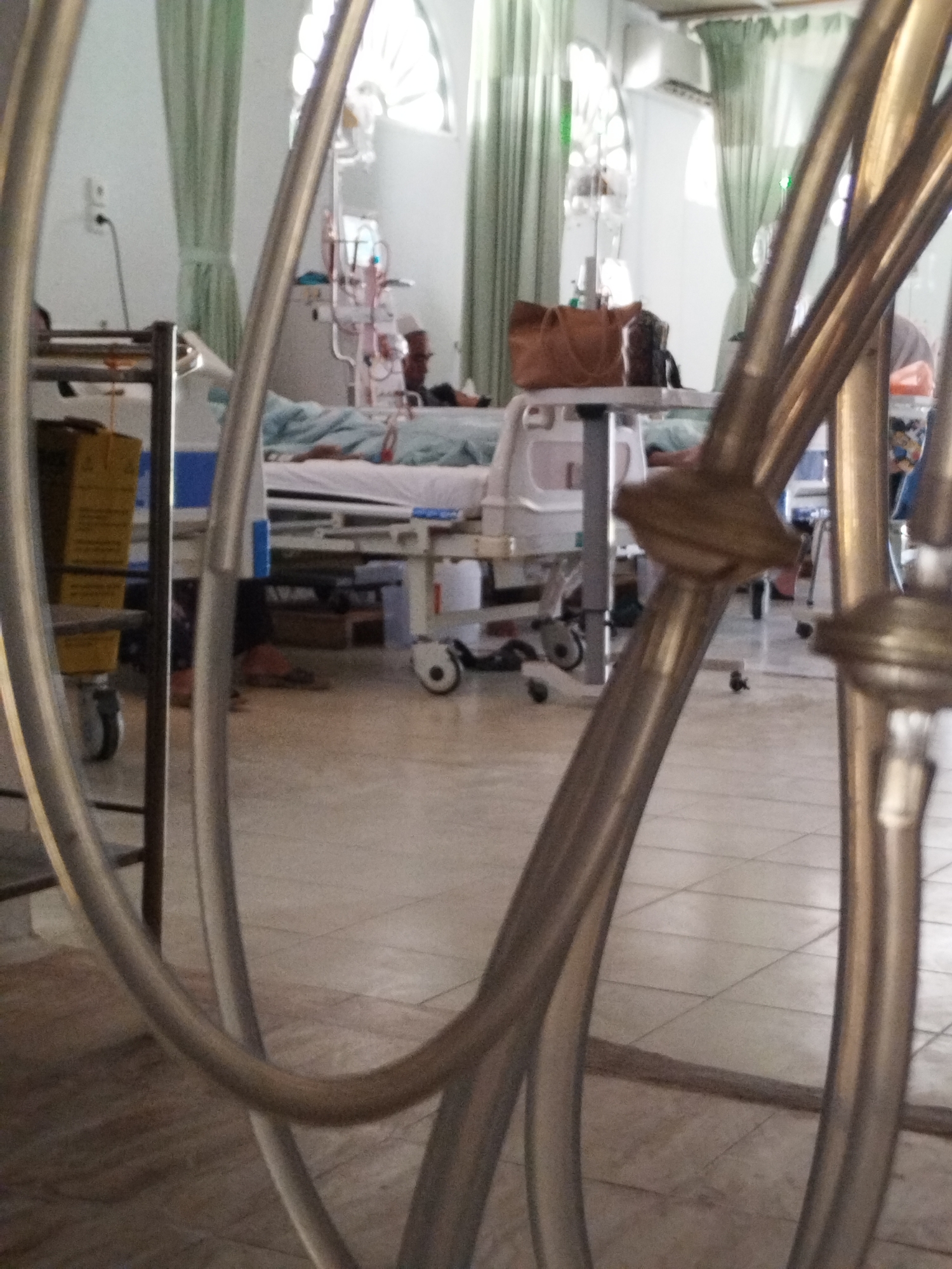 A patient lies in a hospital bed in Lhokseumawe, Indonesia. Visible medical equipment includes a dialysis machine. A visitor sits near the bedside. The room is curtained with semicircular arched windows. The foreground contains decorative metalwork.