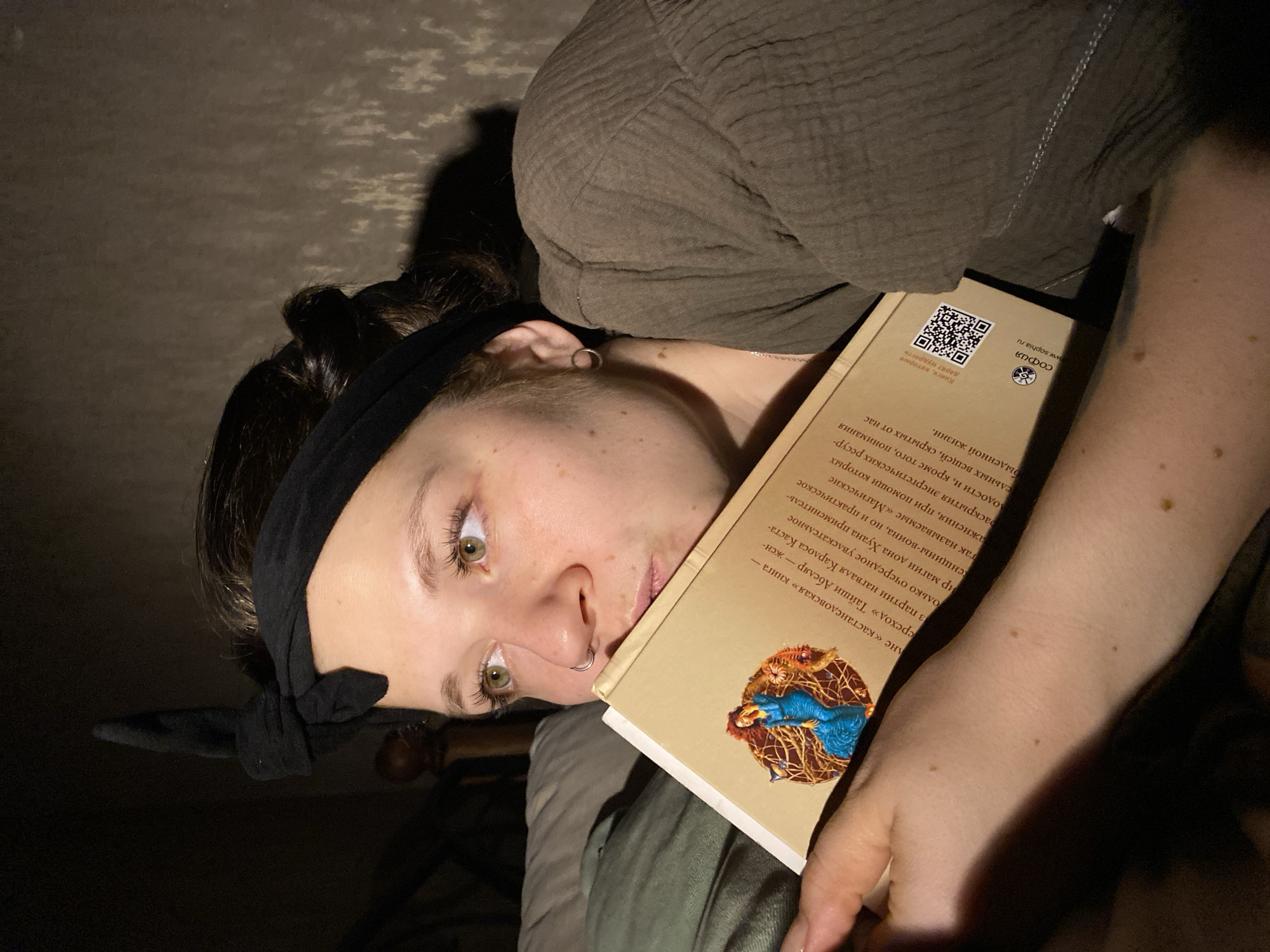 A young woman with a dark headband is holding a book up to her face. The book's cover has a picture of a blue bird. The book text appears to be in Russian. The publisher is "София" (Sophia), and the website is www.sophia.ru. The book description includes "книга Тайи Абеляр" (book Taji Abelar). The book series is "кастанедовская" (Castaneda).
There is no evidence indicating this photo was taken in the city of Chaikovsky, Russia.
