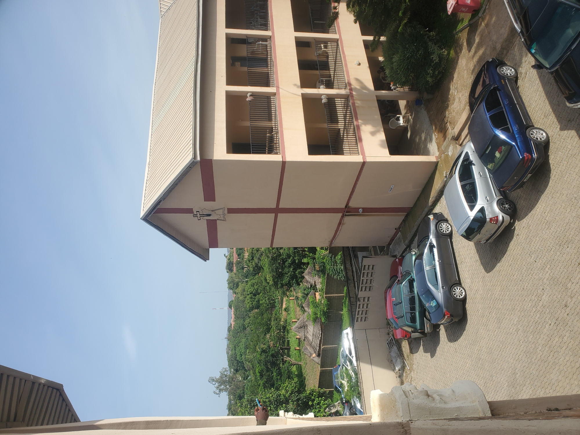 This image is a high-angle, outdoor shot of a building with several cars parked in a courtyard. The building is light tan with maroon trim and appears to be a multi-story structure with balconies. There are also some thatched-roof structures visible in the background, surrounded by lush greenery and trees. The sky is clear and blue. The scene is set in the city of Jos, Nigeria. The courtyard is paved with interlocking bricks. Several cars are parked in various spots. A person's hand is visible on the left side of the frame, holding a blue mobile phone. The overall atmosphere is calm and daytime.