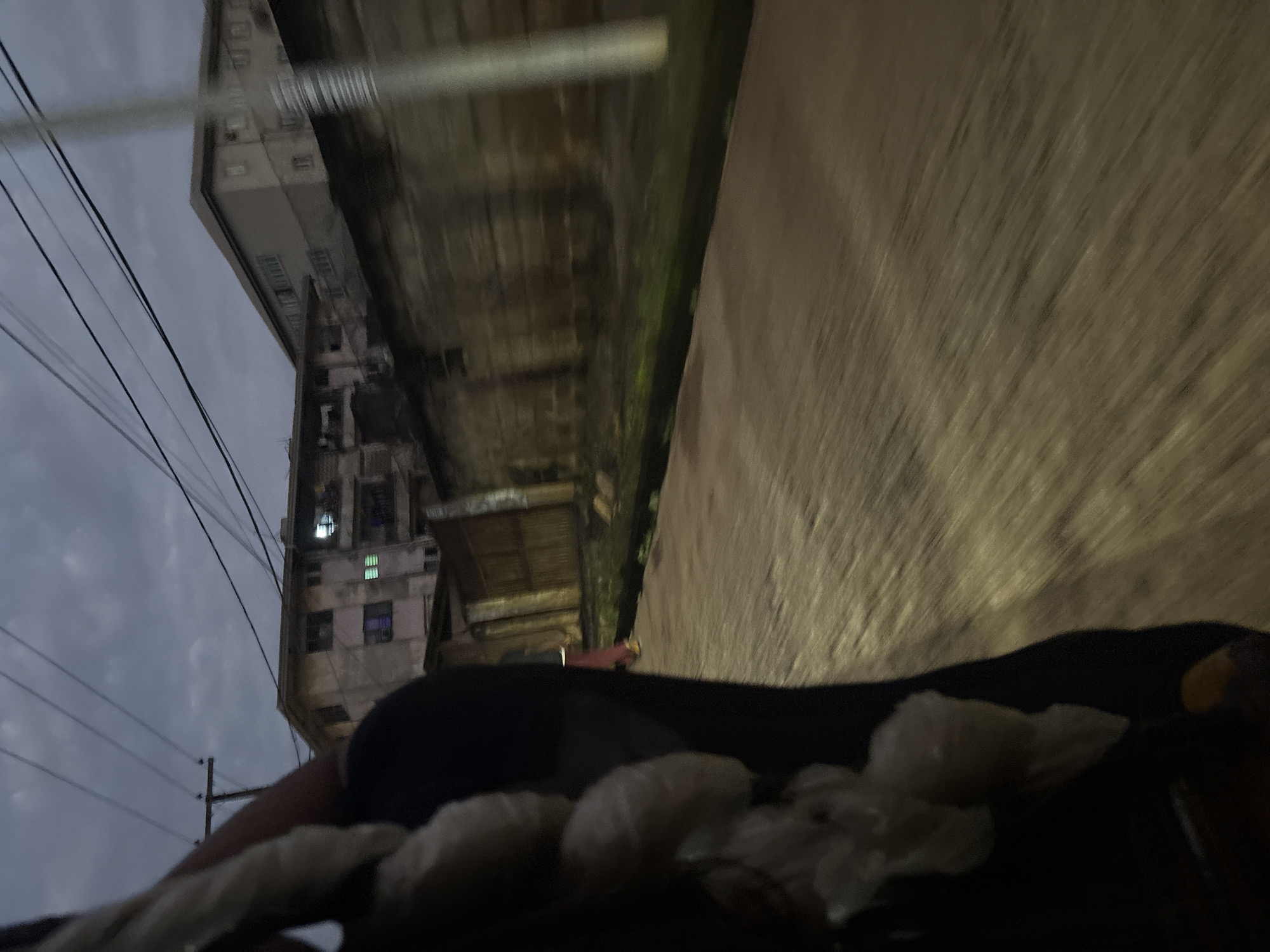The image captures a motion-blurred view of a street scene in Onitsha, Nigeria, likely taken from a moving vehicle. In the foreground, the left side of the frame is dominated by a dark, blurry mass, possibly clothing or a person's arm, with a string of pale, translucent objects hanging from it. The background features several multi-story buildings with visible windows, some illuminated, suggesting it is either dusk or night. A textured, weathered wall runs along the middle ground. Power lines are visible crisscrossing the cloudy sky. The ground appears to be paved with a rough texture, and a narrow strip of green vegetation is visible along the base of the wall.