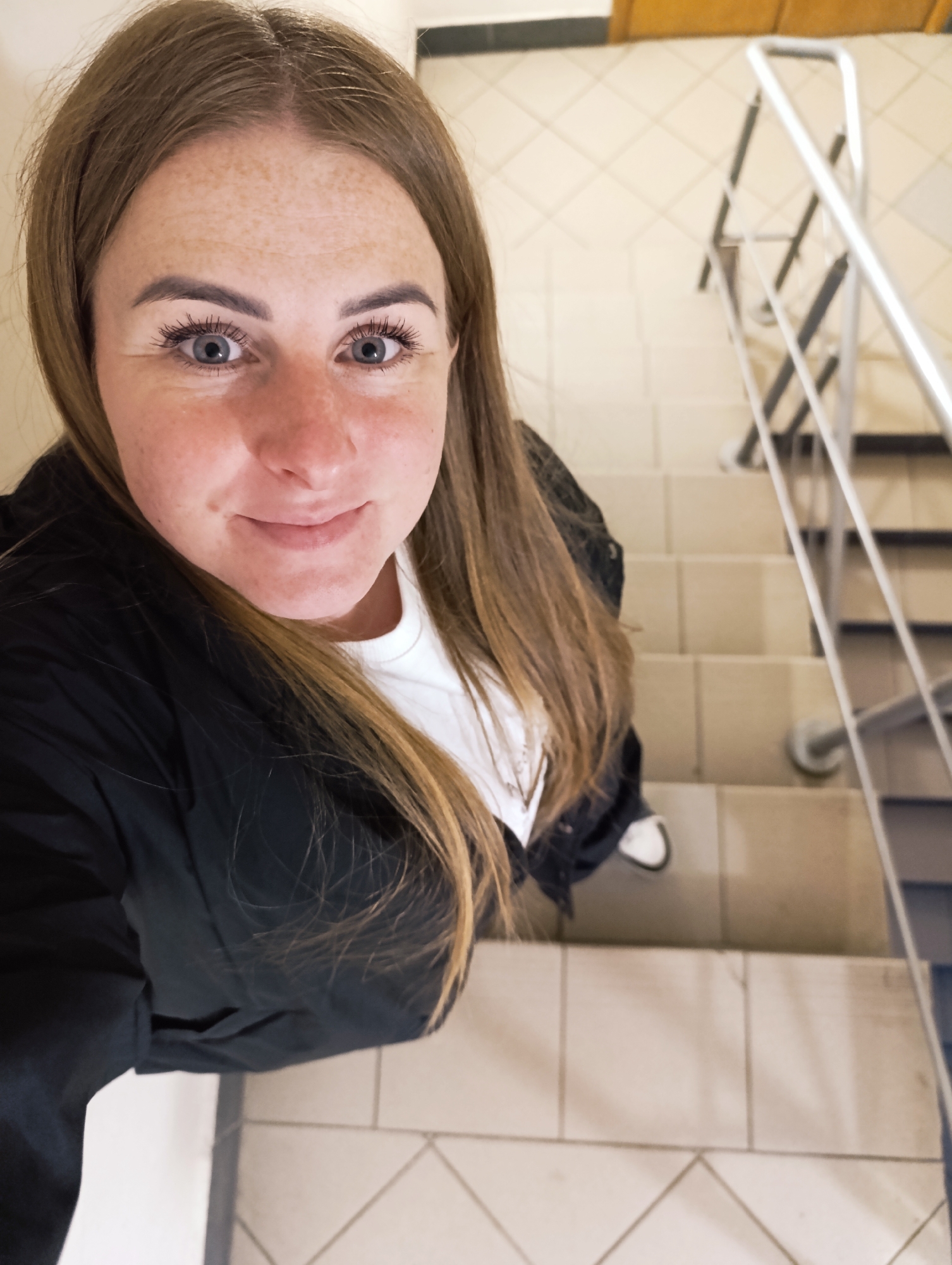 A fair-skinned woman with shoulder-length light brown hair stands on a staircase, looking up at the camera. She has freckles on her face and is wearing mascara on her eyelashes. She is dressed in a white t-shirt underneath a black jacket. A partial view of a metal railing is visible on the right side of the image. The staircase is tiled. The location appears to be inside a building, city and state Unknown, Unknown.