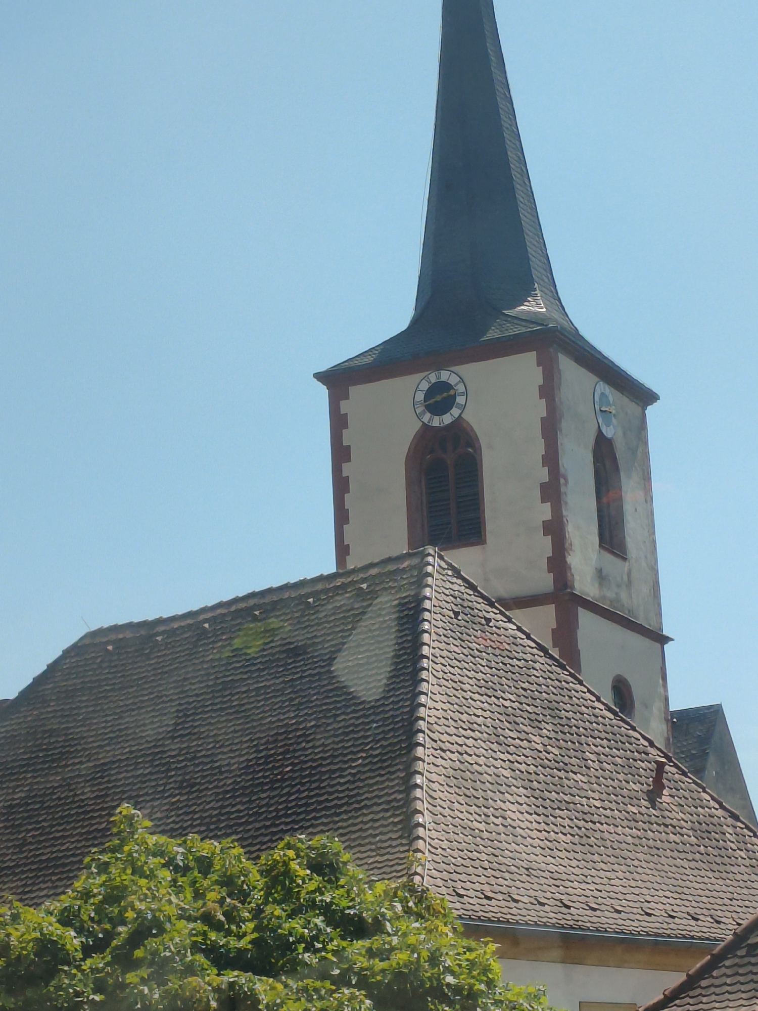 The image shows the spire of a church in Ochsenfurt, Germany. The church has a white tower with red trim and two clocks on its facade. The spire is dark and pointed. In the foreground, there is a tiled roof and some green trees. The sky is clear and blue.