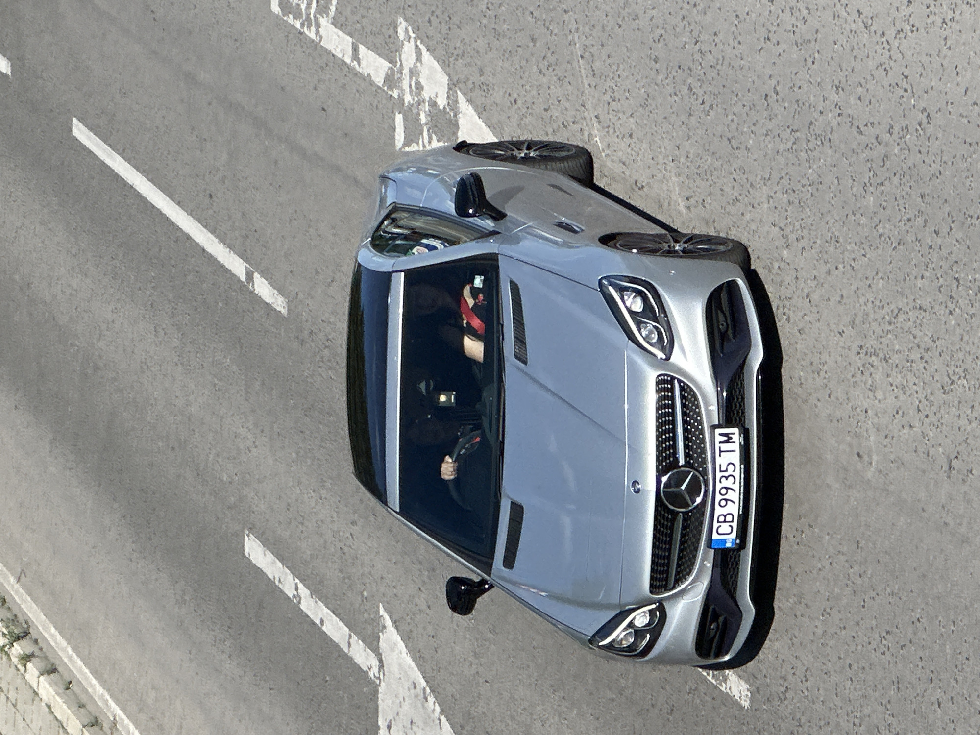 The image shows a silver Mercedes-Benz SLK roadster driving on a road in Veliko Tarnovo, Bulgaria. The car's license plate reads "CB 9935 TM". There are two people inside the car, a driver and a passenger. The driver is wearing a dark jacket and the passenger is wearing a red shirt. The car is on a multi-lane road with white lane markings. The road appears to be asphalt. The surrounding environment is not clearly visible but appears to be urban or suburban.