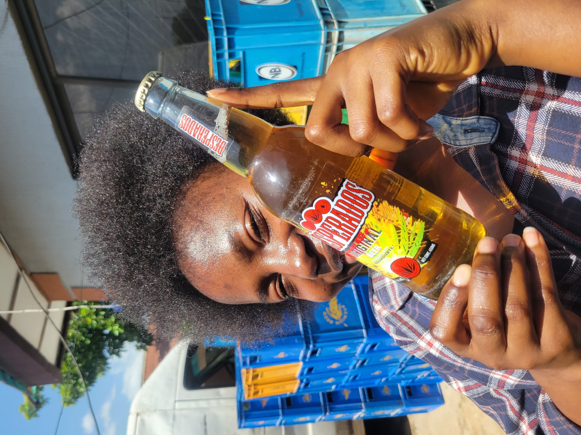 A young African woman with dark, curly hair is holding a bottle of Desperados beer in front of her face, which is partially obscured by the bottle. She is wearing a plaid shirt. The background shows stacks of blue and yellow plastic crates, a white vehicle, and a building with a brown roof. The scene appears to be outdoors, possibly in an urban setting like Jos, Nigeria, given the presence of crates often used for beverages and the style of the building. The lighting suggests it is daytime.