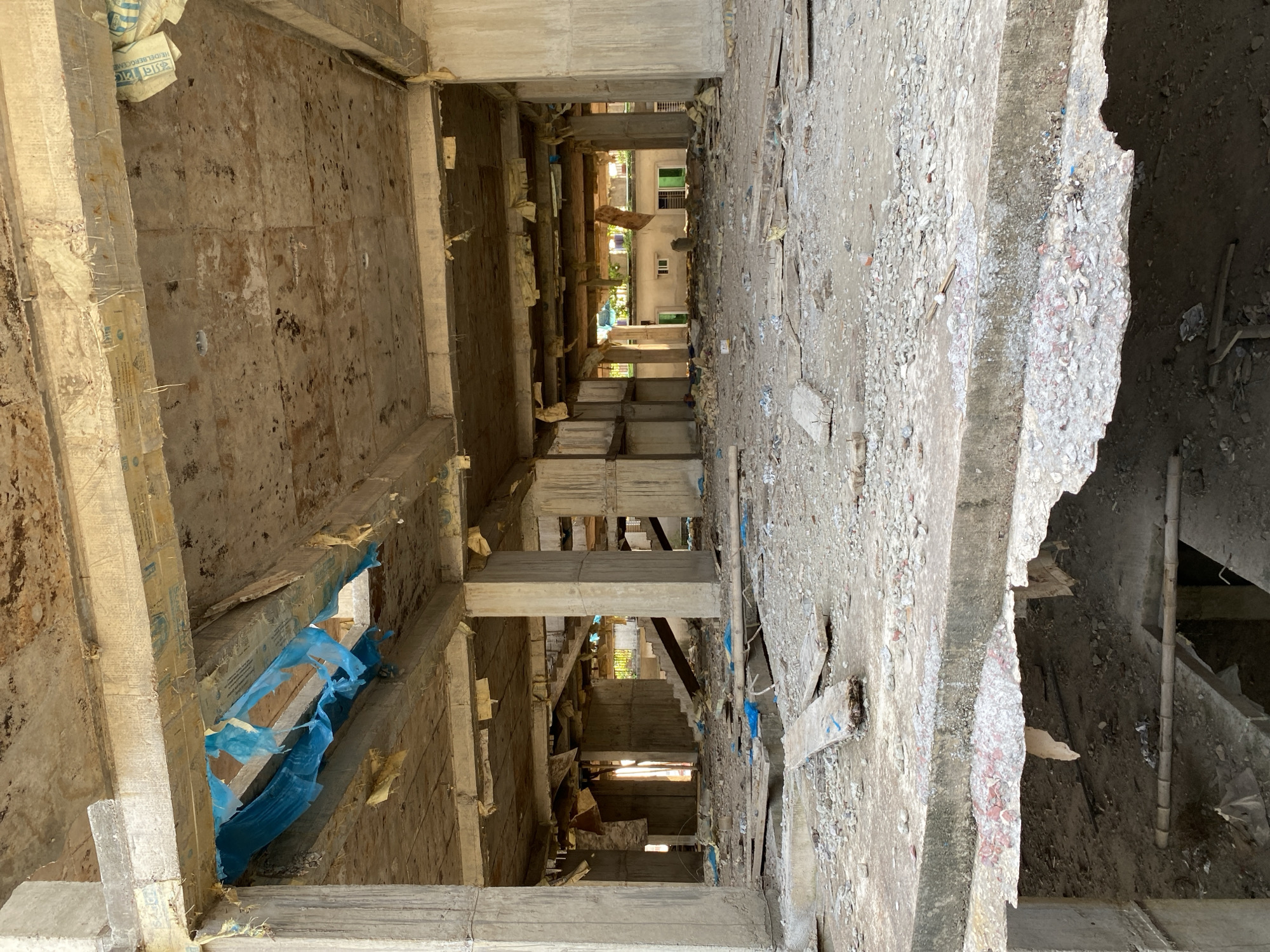 The image shows the interior of an unfinished building, likely a residential or commercial structure, under construction in Dhaka, Bangladesh. The view is a vertical shot looking down through the floors, with exposed concrete walls, pillars, and staircases. The walls show signs of wear and debris from construction. Construction materials such as blue plastic sheets and wooden supports are visible. The distant view shows completed buildings outside, suggesting the location is in an urban area. No people are visible. The scene conveys a sense of abandonment or unfinished construction.