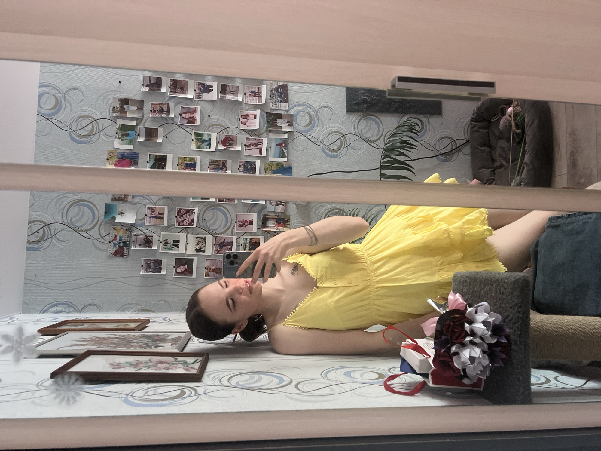 The image depicts an indoor scene in Chaikovsky, Russia, captured as a mirror selfie. A young female with brown hair tied back and wearing a yellow strappy, tiered dress with white trim is visible. She leans against a light blue wallpapered wall adorned with swirling patterns. Her left hand holds a dark-colored smartphone, used to take the photograph, and she has several thin bracelets on her left wrist, a small hoop earring in her left ear, and a tattoo on her upper left arm. To her left, on a grey carpeted cat tree platform, rests a small white gift bag with red handles, an open white box, and a bouquet of origami-style flowers in red, white, and purple. The wall behind her also displays two framed pictures and two white, snowflake-like decorations. Reflected in the mirror to her right, the wall is covered with a collage of numerous small photographs pinned and strung together. Further in the reflection, a dark grey, circular pet bed containing small toys is on the floor, with a large green leafy plant partially visible behind it.