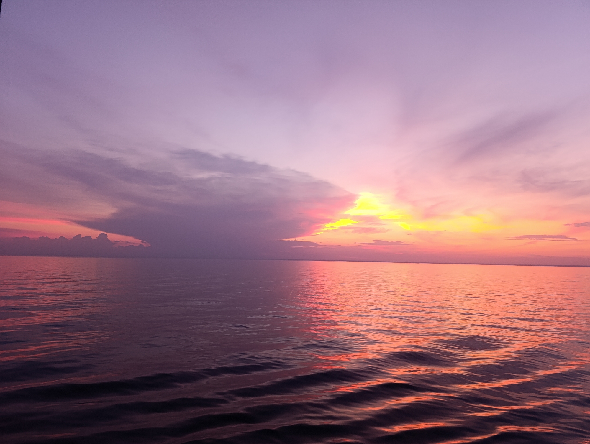 The image shows a wide body of water, likely a lake or ocean, reflecting the vibrant colors of a sunset. The sky is a gradient of purple, pink, and bright yellow, with rays of light emanating from behind a cloud formation on the right side of the frame. The water's surface is rippled, and it mirrors the sky's colors, creating a dual-toned effect with the left side appearing darker and more purplish, while the right side is awash in pink and orange hues. There are no people, structures, or specific landmarks visible that would indicate a precise location. The scene is devoid of any human activity or man-made objects, presenting a purely natural landscape.