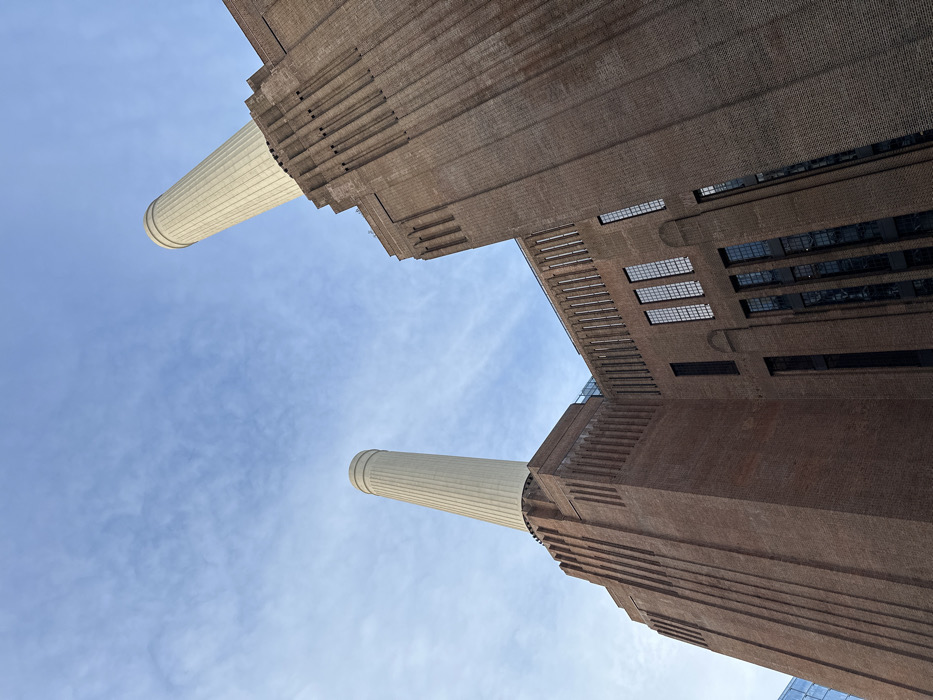 Looking up at Battersea Power Station, two tall, light-colored chimneys dominate the view against a pale blue sky with wispy clouds. The brick architecture of the station is visible, featuring layered, stepped patterns and rows of rectangular windows. The perspective emphasizes the height and massiveness of the building.